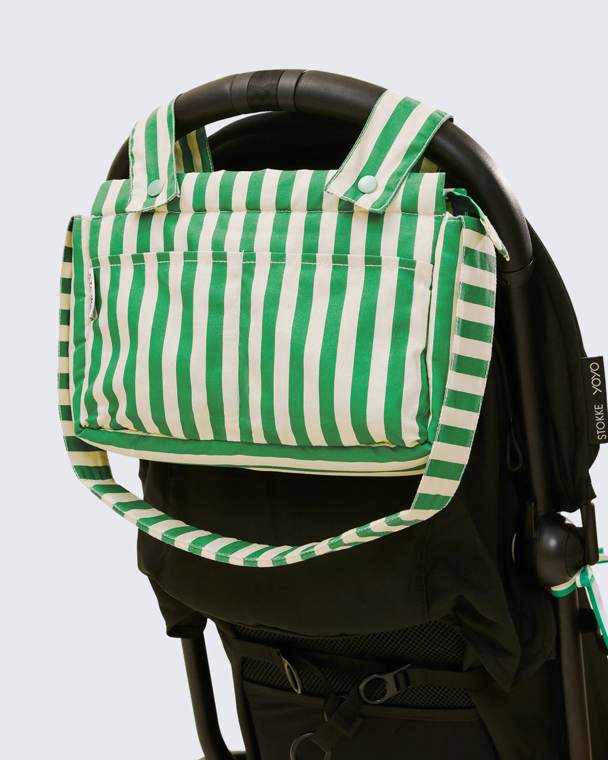 Black stroller with Stroller Party™ Caddy Bag in Green Stripe hanging off the back handlebar, on a beige background.