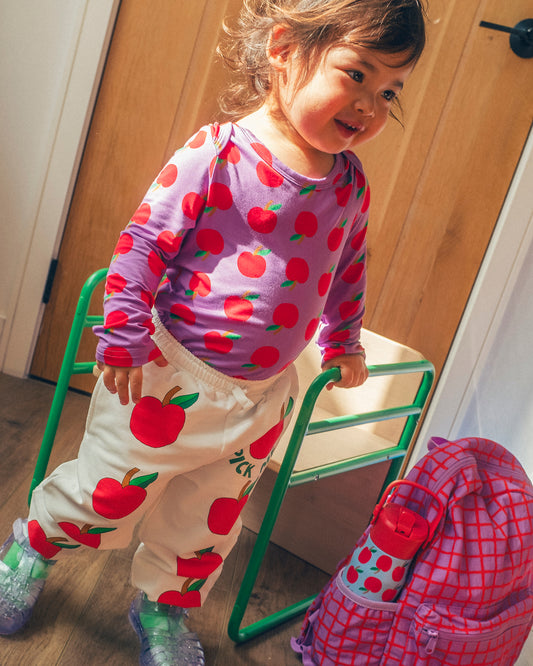 A toddler wearing Long Sleeve Bamboo Onesie Two-Pack in Apple and French Terry Jogger in Ivory Apple, standing next to a green metal chair with Cool Kid Backpack in Orchid Grid.