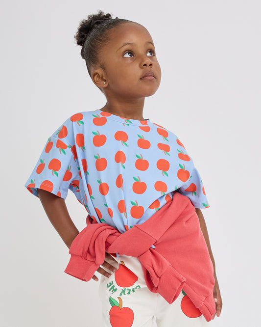 A young girl with her hair in a bun is wearing Graphic Tee in Blue Apple, and French Terry Jogger in Ivory Apple. She is looking up and to the side against a light gray studio background.