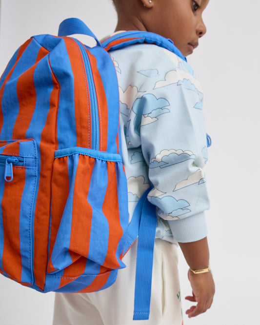 A child wearing Graphic Sweatshirt in Blue Clouds, shown from the side/back wearing a Cool Kid Backpack in Blue & Rust Stripe against a plain light gray background.