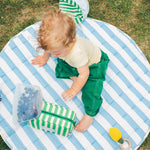 Toddler sitting outside on Inside/Outside Play Mat in Blue Wide Stripe, reaching for Mini Cooler Bag in Green Stripe, surrounded by a bag of blueberries and chewy toy with the Big Floppy Sun Hat in Blue Pinstripe on the grass.