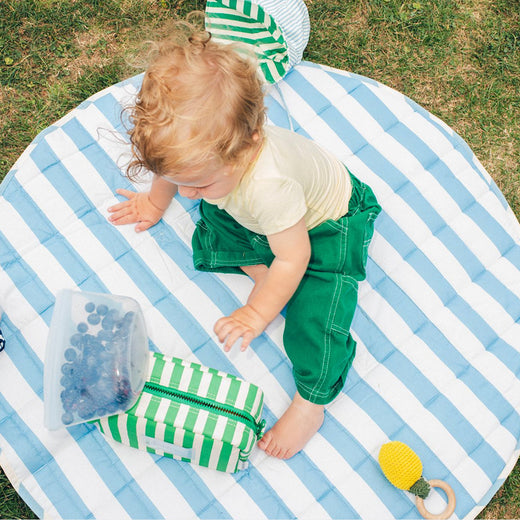 Toddler sitting outside on Inside/Outside Play Mat in Blue Wide Stripe, reaching for Mini Cooler Bag in Green Stripe, surrounded by a bag of blueberries and chewy toy with the Big Floppy Sun Hat in Blue Pinstripe on the grass.