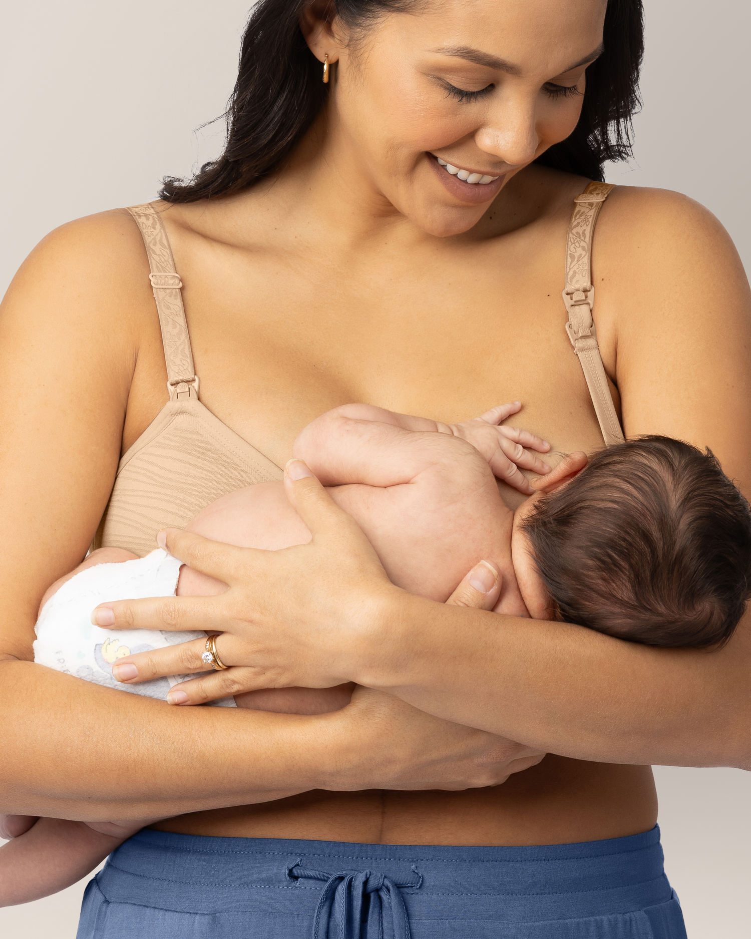 Woman wearing beige hands-free pumping & nursing bra, holding and nursing baby