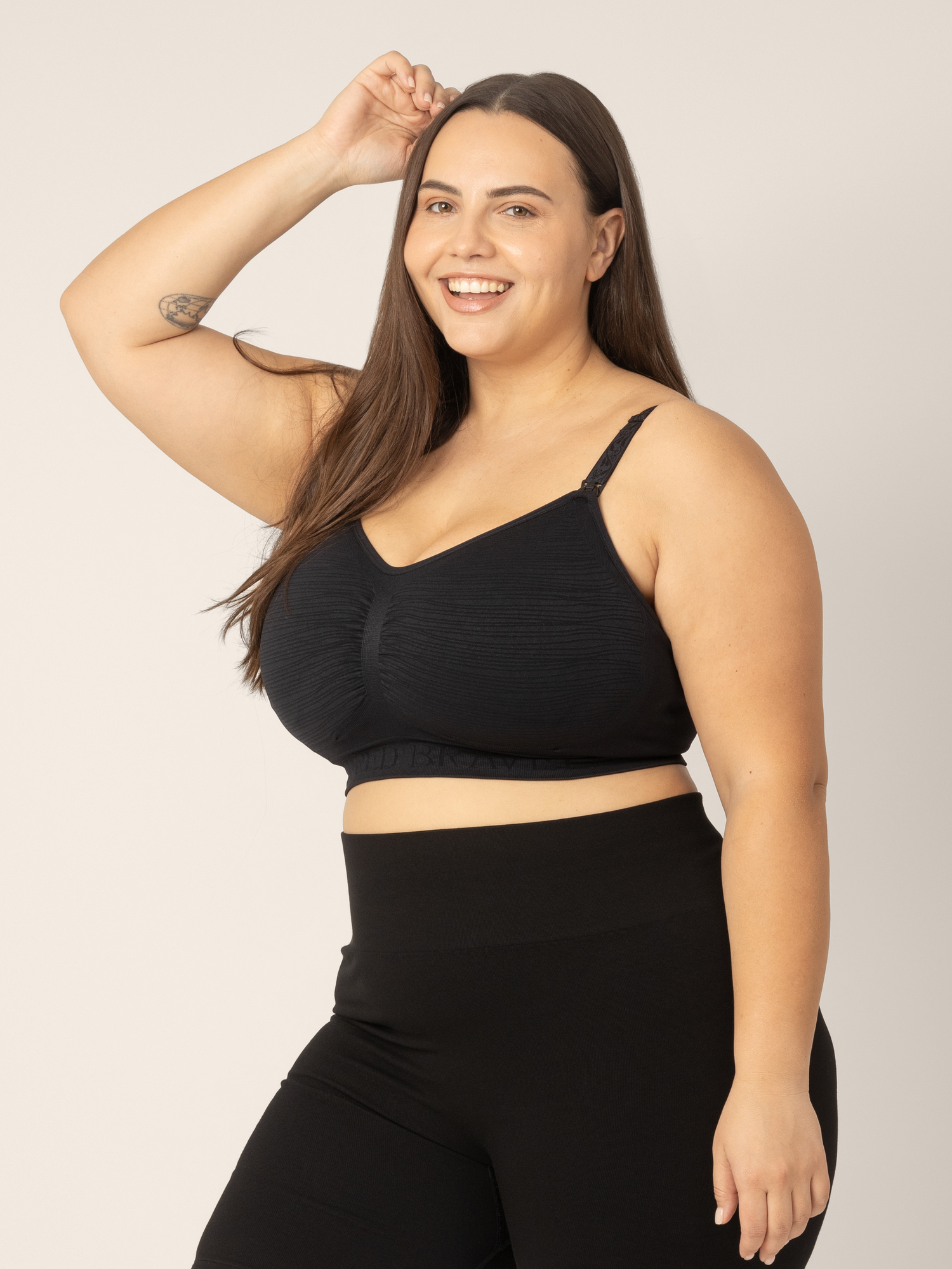 Side view of super busty model wearing the Sublime Hands-Free Pumping & Nursing Bra in Black. Her arm is up and hand over head showing the movement of the bra. @model_info:Anna is wearing a Large Super Busty.