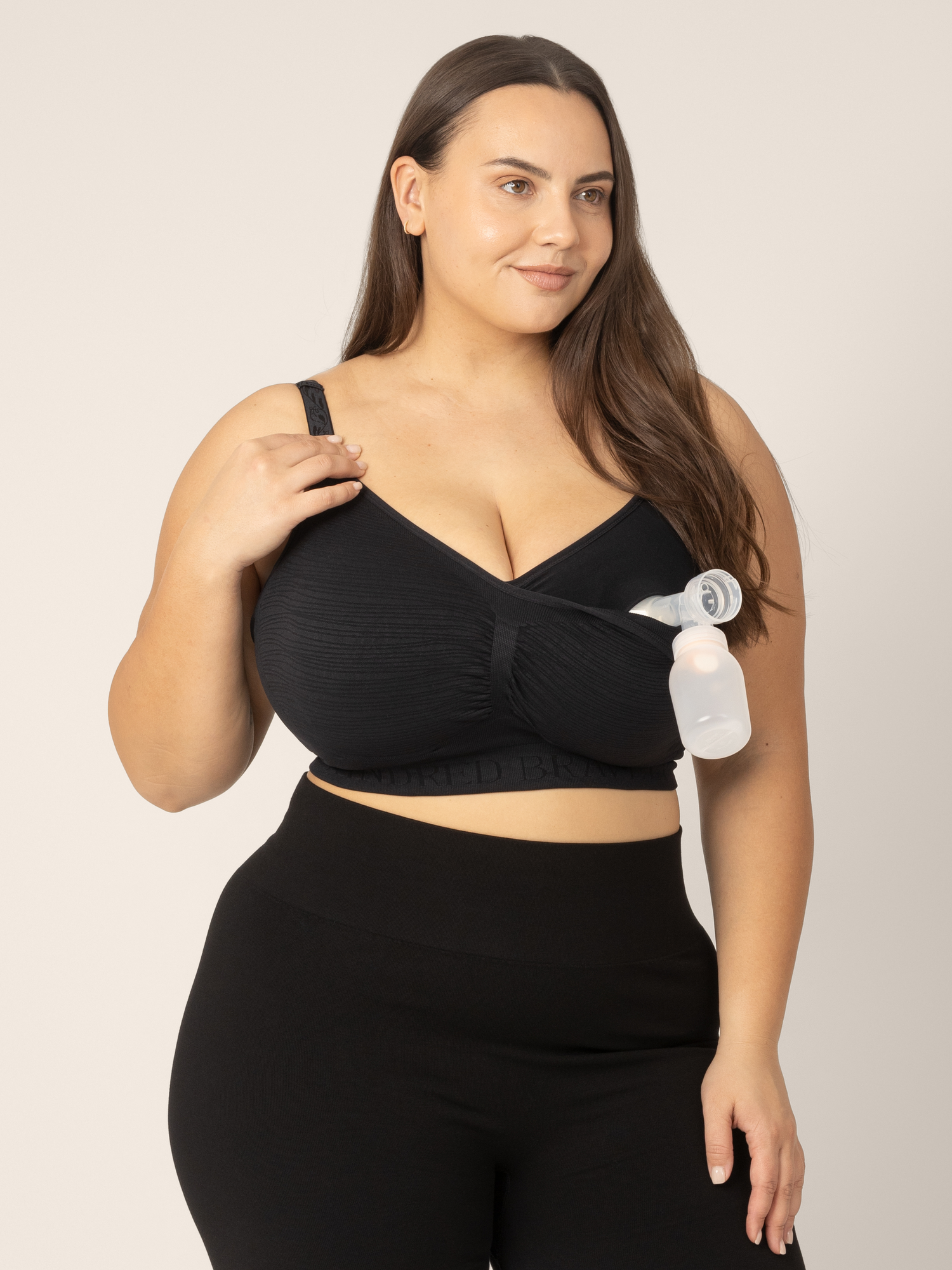 Super busty model wearing the Sublime Hands-Free Pumping & Nursing Bra in Black. The pumping layer is clipped down to show the bra pump inserted, for hands-free pumping access.
