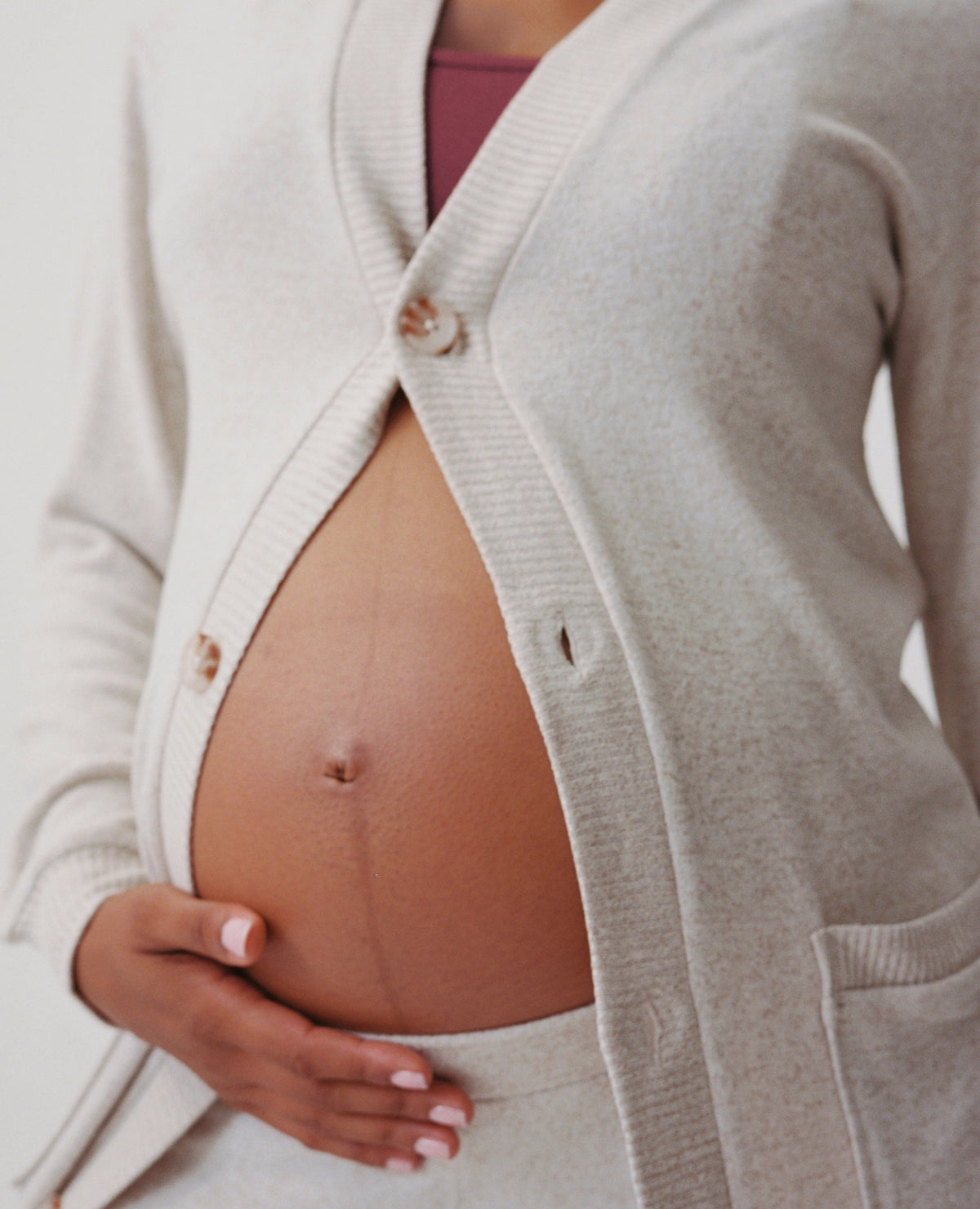 Close up of model wearing CozyKnit Wide Leg Lounge Pant in Oat, and CozyKnit Long Cardigan in Oat, and French Terry Maternity & Nursing Bralette in Deep Burgundy on a beige background.