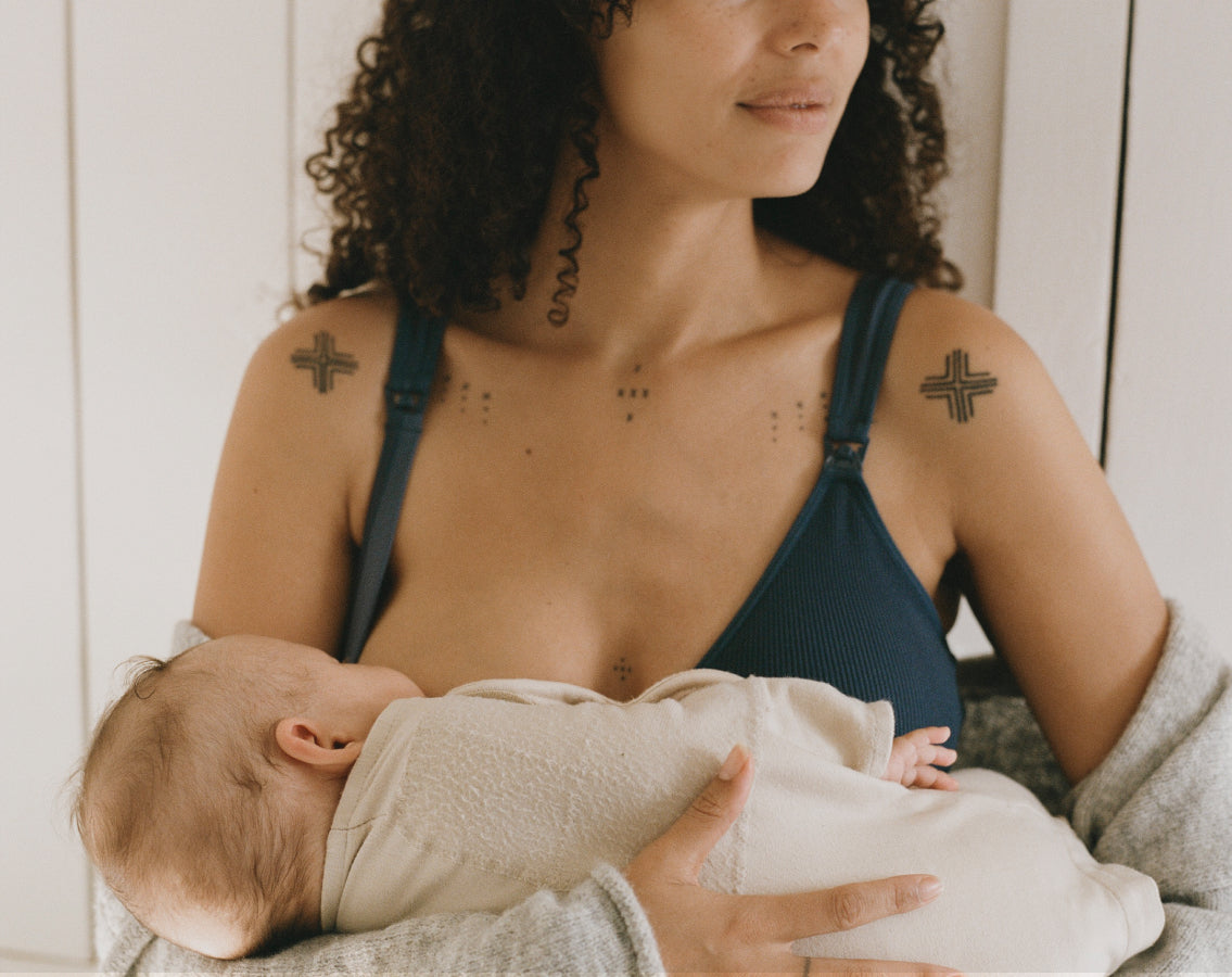 Model wearing Mia Maternity & Nursing Lounge Bra in Light Navy while a baby nurses, in light filled room and window blinds in background.