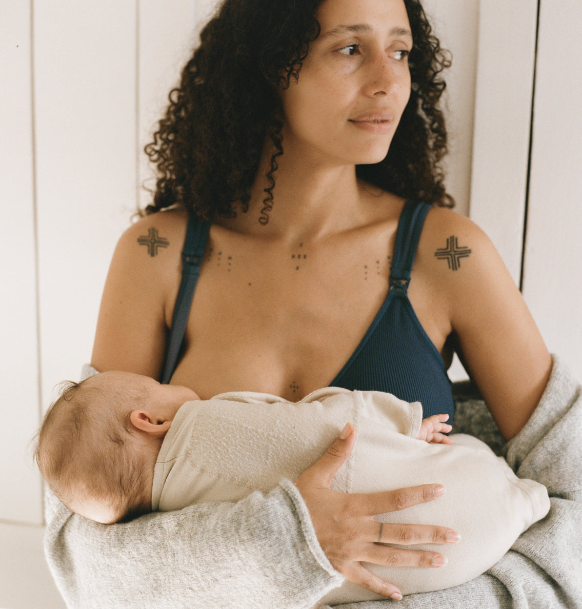 Model wearing Mia Maternity & Nursing Lounge Bra in Light Navy while a baby nurses, in light filled room and window blinds in background.