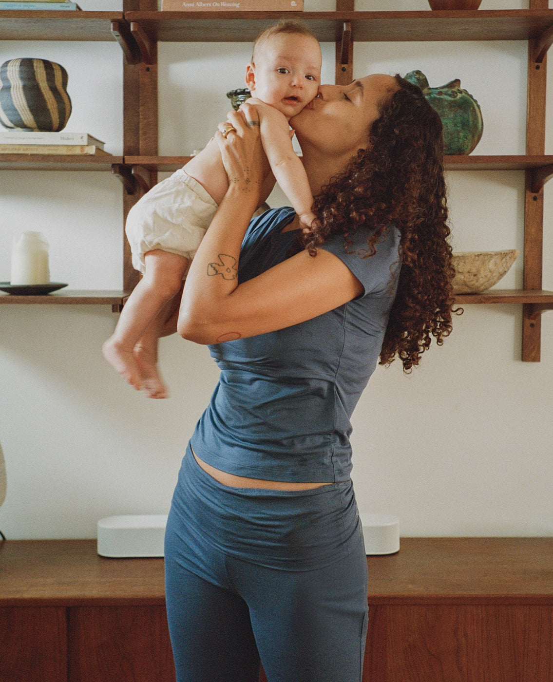 A woman in The Sunday Maternity & Nursing Pajama Set in Slate Blue holds a baby close to her face in a warm indoor setting with wooden shelves in the background.