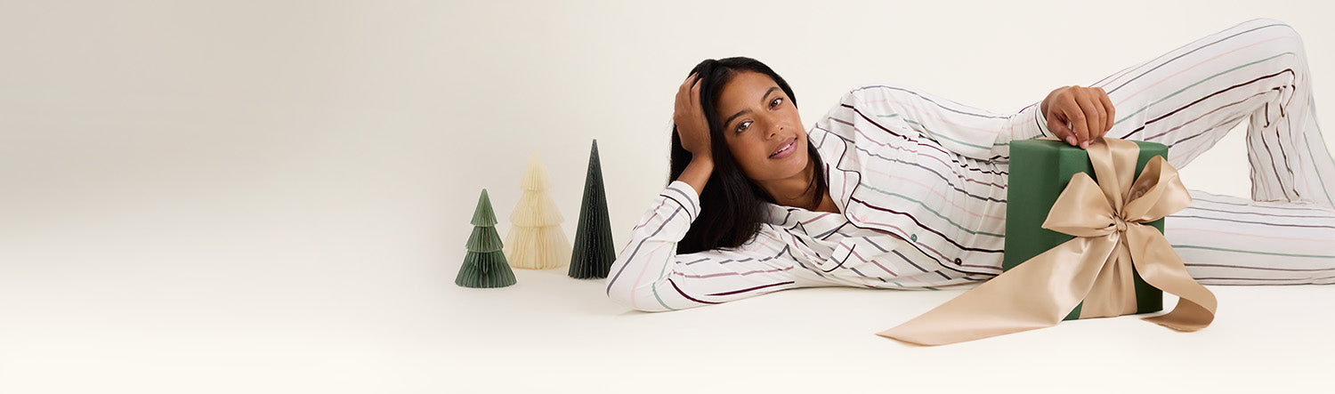 Woman laying on floor next to a gift and decorative paper trees, wearing the Clea Bamboo Long Sleeve Pajama Set in Classic Stripe