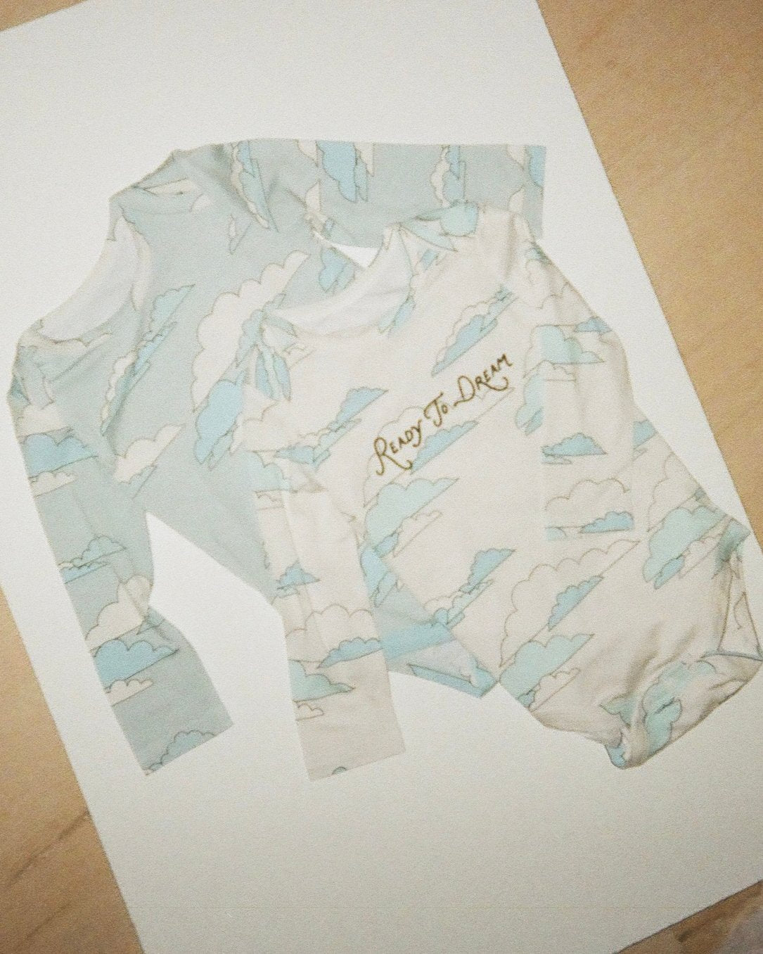 Stylized image of Long Sleeve Bamboo Onesie Two-Pack in Clouds on a white sheet on a wooden floor. 