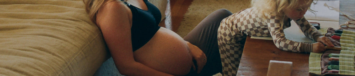 A pregnant woman sits on the floor against a beige sofa next to a young child at a wooden table playing with art materials and wearing Bamboo Baby Two-Piece Pajama Set in Taupe Checker.