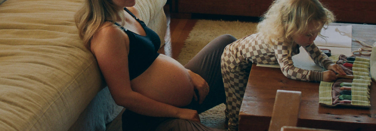 A pregnant woman sits on the floor against a beige sofa next to a young child at a wooden table playing with art materials and wearing Bamboo Baby Two-Piece Pajama Set in Taupe Checker.