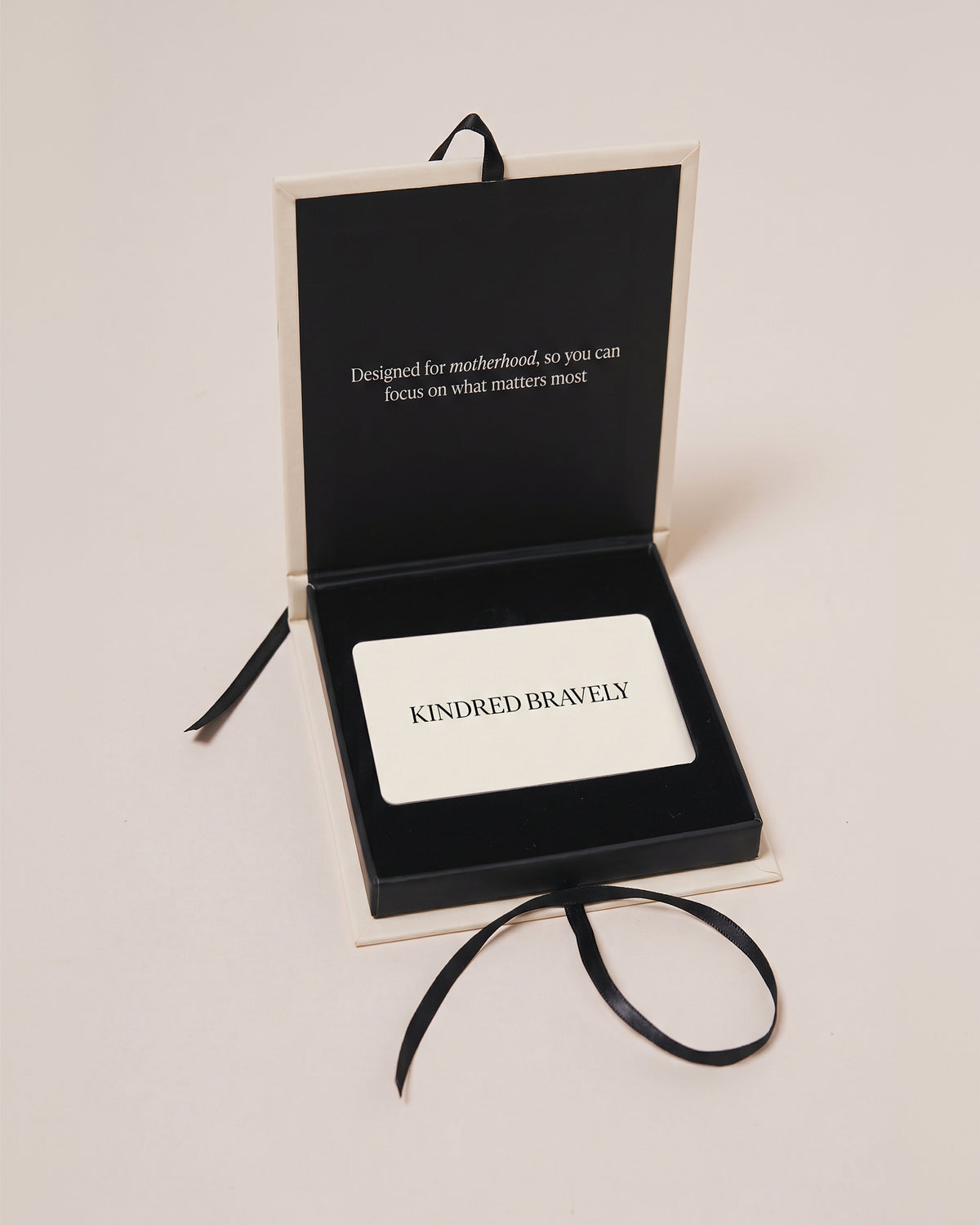An open black presentation box with cream-colored edges on a light beige background. The box contains a white card printed with the word 'KINDRED BRAVELY' in black capital letters, and white text that reads, Designed for motherhood, so you can focus on what matters most.