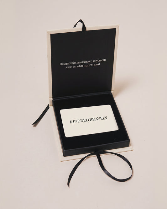 An open black presentation box with cream-colored edges on a light beige background. The box contains a white card printed with the word 'KINDRED BRAVELY' in black capital letters, and white text that reads, Designed for motherhood, so you can focus on what matters most.