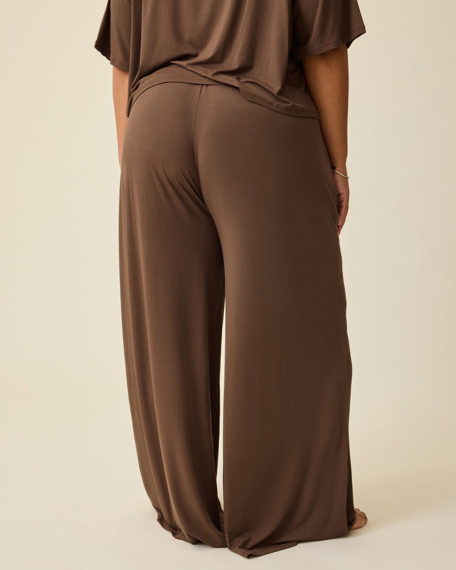 Back view of model wearing Aria Bamboo Casual Cropped Short Sleeve Top in Chocolate and Aria Bamboo Casual Lounge Pant in Chocolate, on a beige background. 