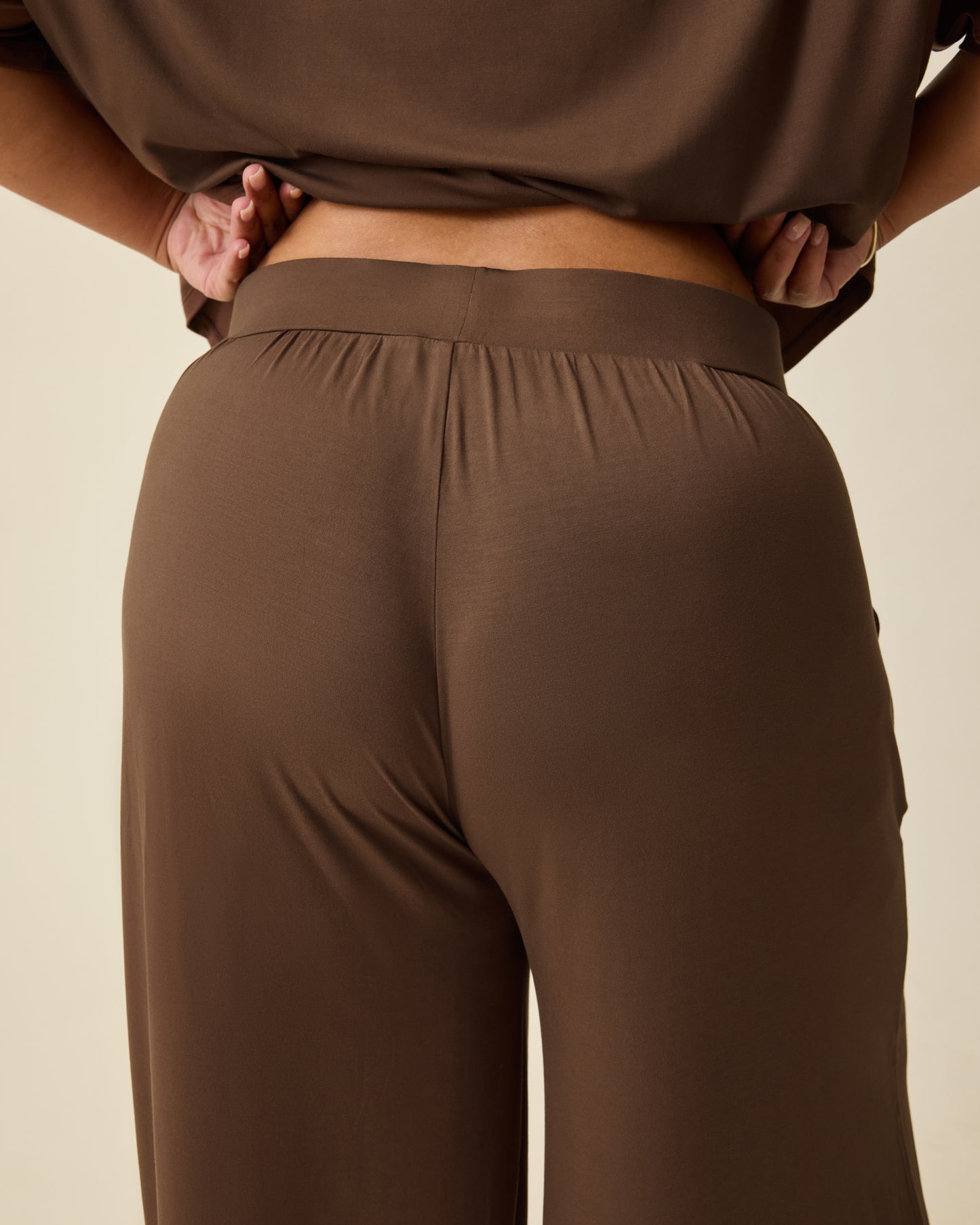 Back view of model wearing Aria Bamboo Casual Cropped Short Sleeve Top in Chocolate and Aria Bamboo Casual Lounge Pant in Chocolate, on a beige background. 