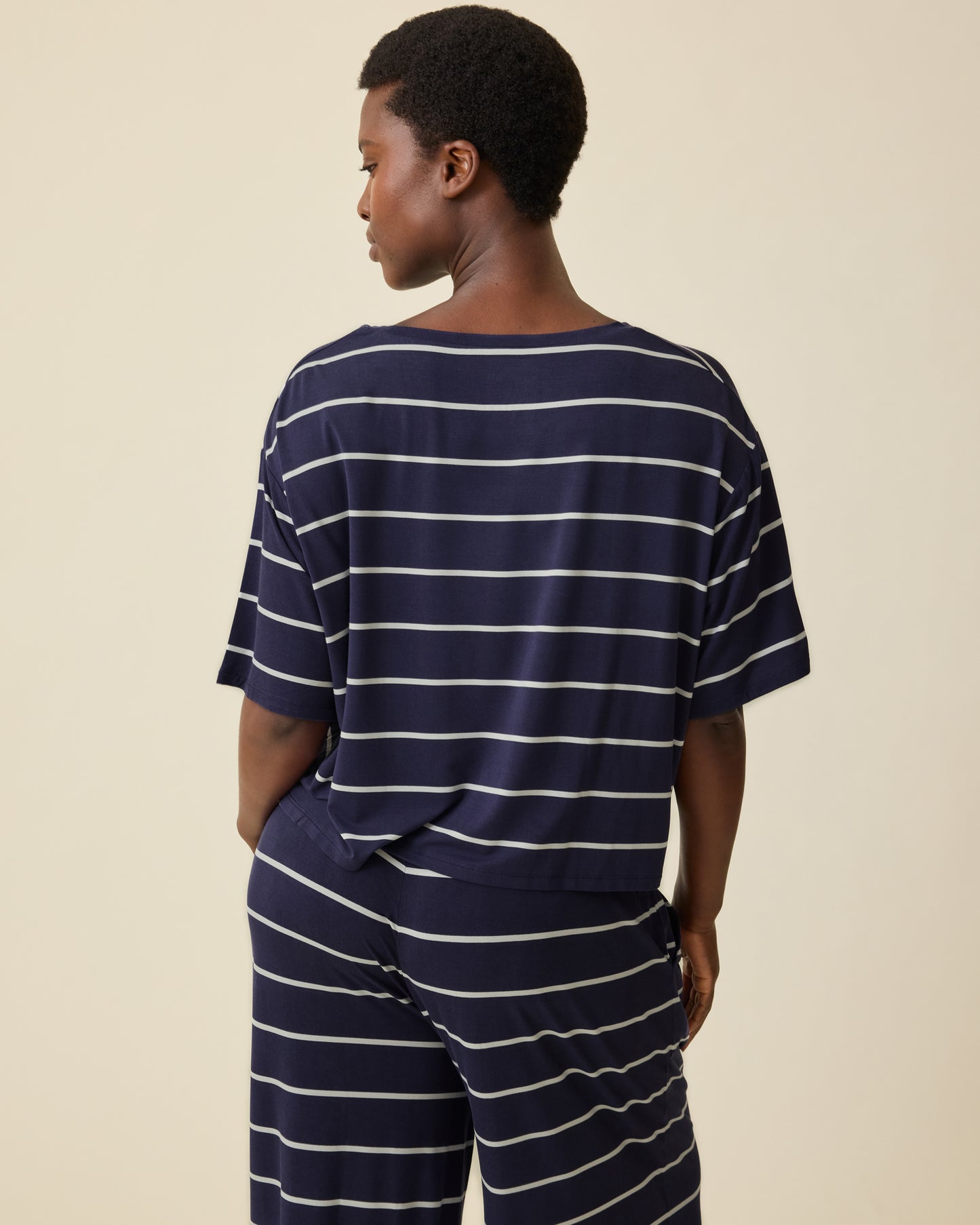 Back view of the Model wearing Aria Bamboo Casual Cropped Short Sleeve Top in Navy & White Stripe and Aria Bamboo Casual Lounge Pant in Navy & White Stripe on a beige background.