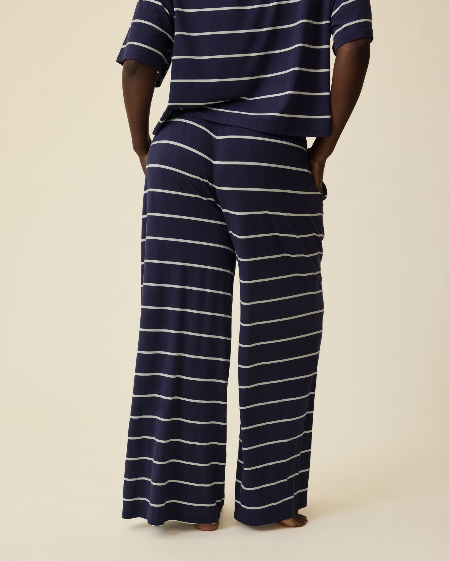 Back view of Model wearing Aria Bamboo Casual Cropped Short Sleeve Top in Navy & White Stripe and Aria Bamboo Casual Lounge Pant in Navy & White Stripe on a beige background.