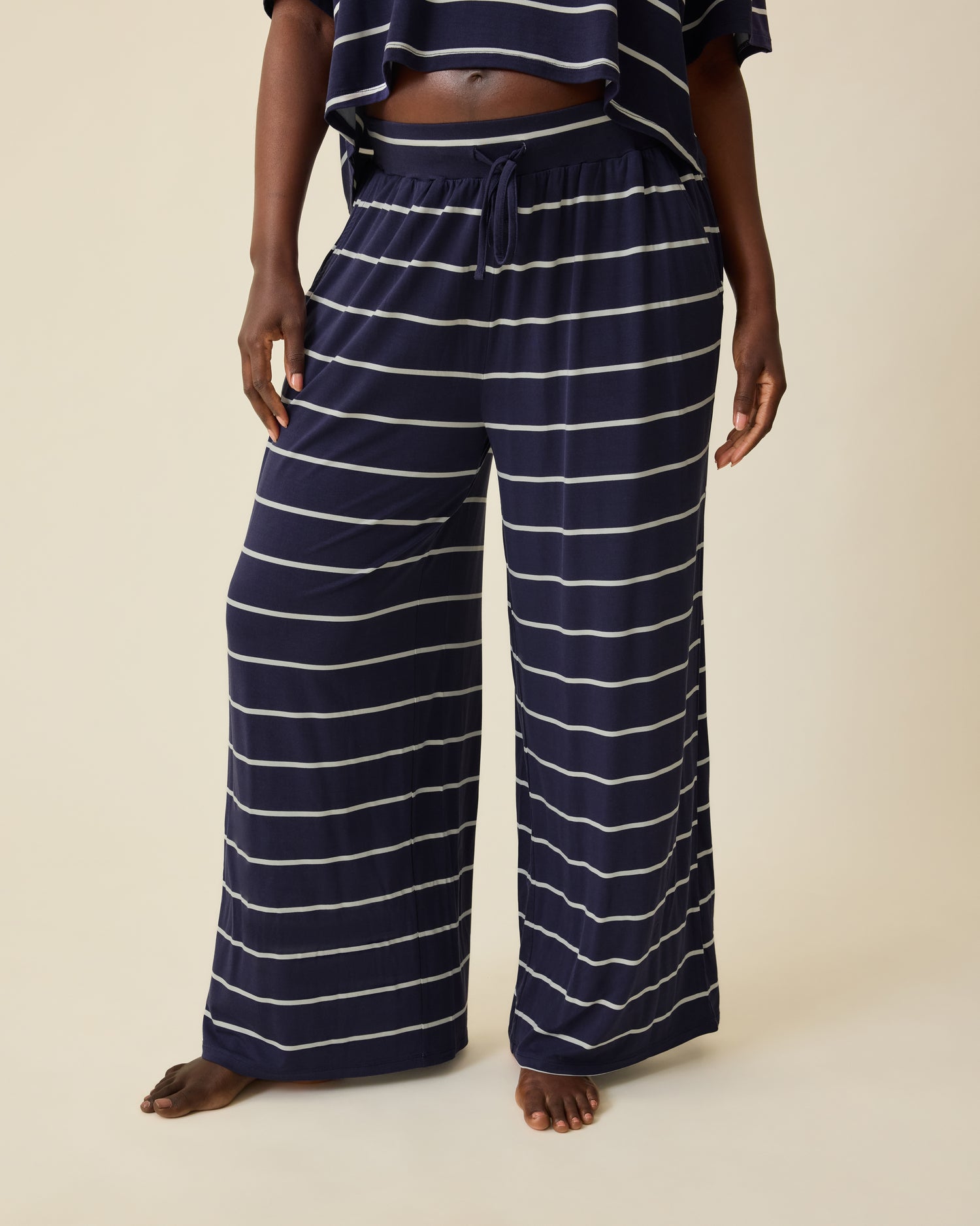 Model is wearing Aria Bamboo Casual Cropped Short Sleeve Top in Navy & White Stripe and Aria Bamboo Casual Lounge Pant in Navy & White Stripe on a beige background. @model_info:Anna is 5'8.5" and wearing a Medium.