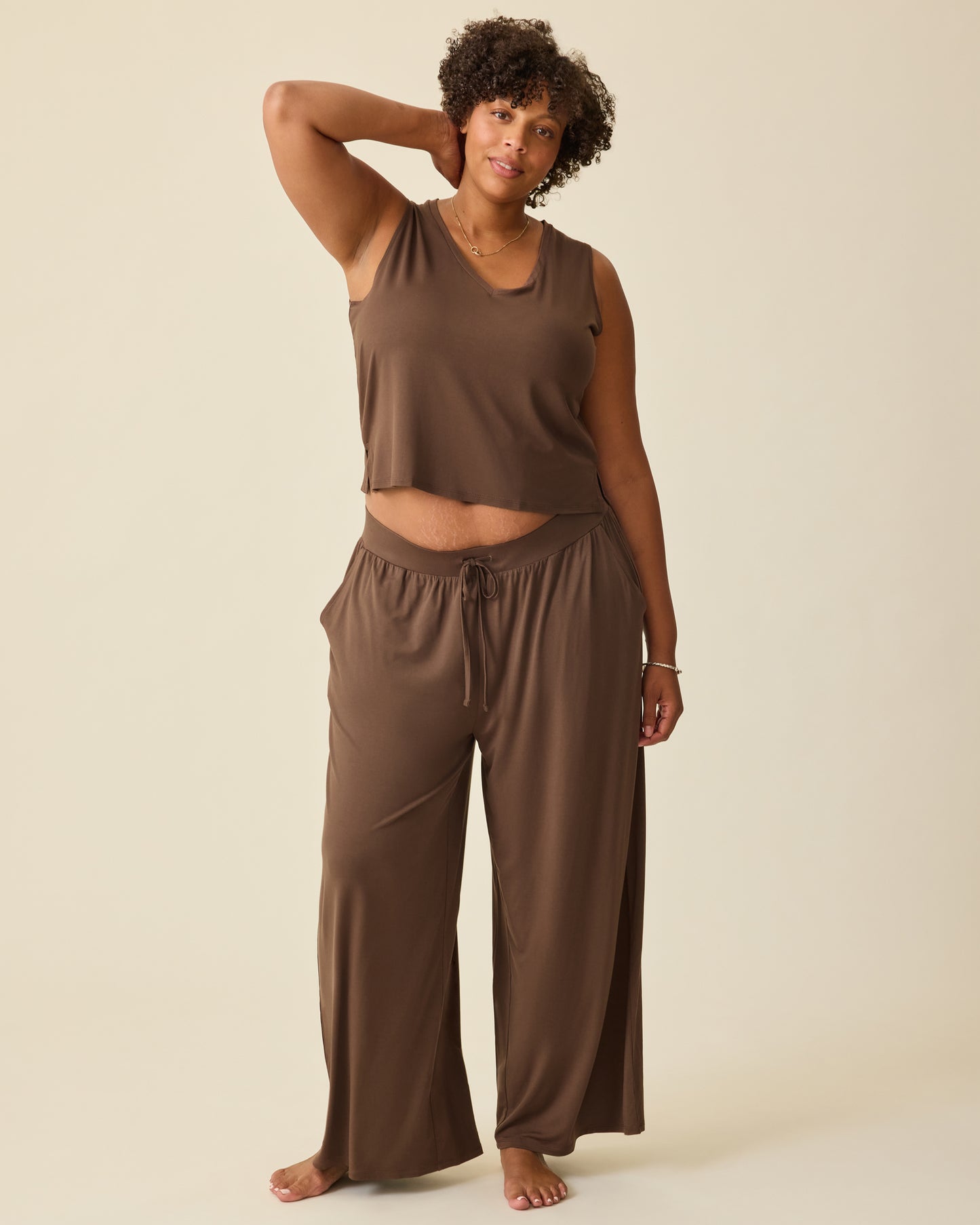 Model is wearing Aria Bamboo Cropped V-neck Tank in Chocolate and Aria Bamboo Casual Lounge Pant in Chocolate on a beige backgound.
