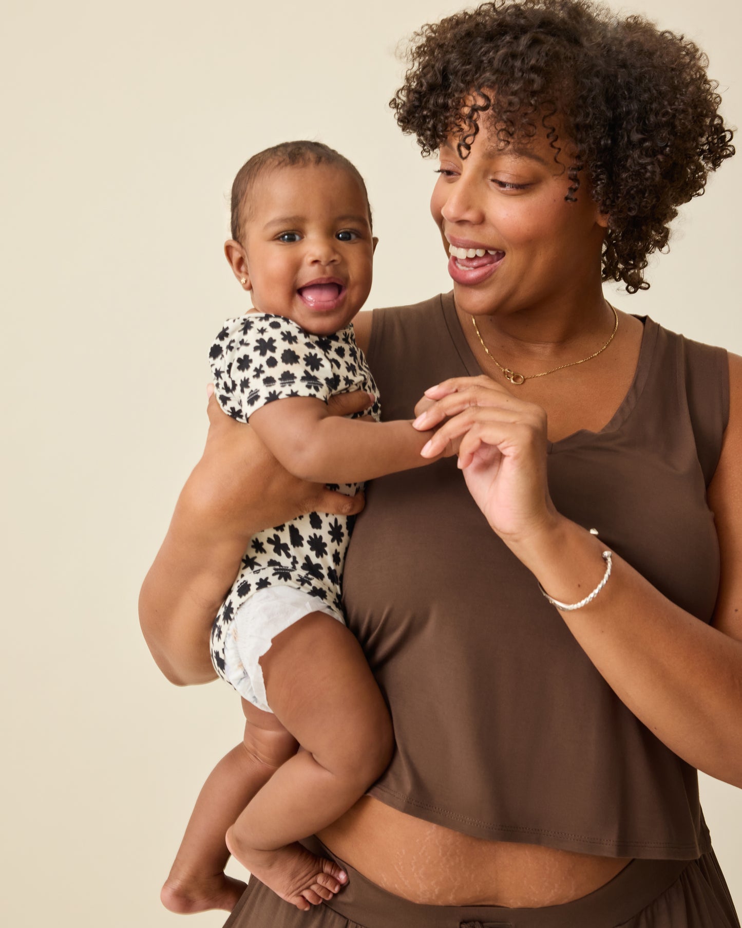 Model is holding baby in Cream Chamomiles Onesie and wearing Aria Bamboo Cropped V-neck Tank in Chocolate and Aria Bamboo Casual Lounge Pant in Chocolate on a beige background.