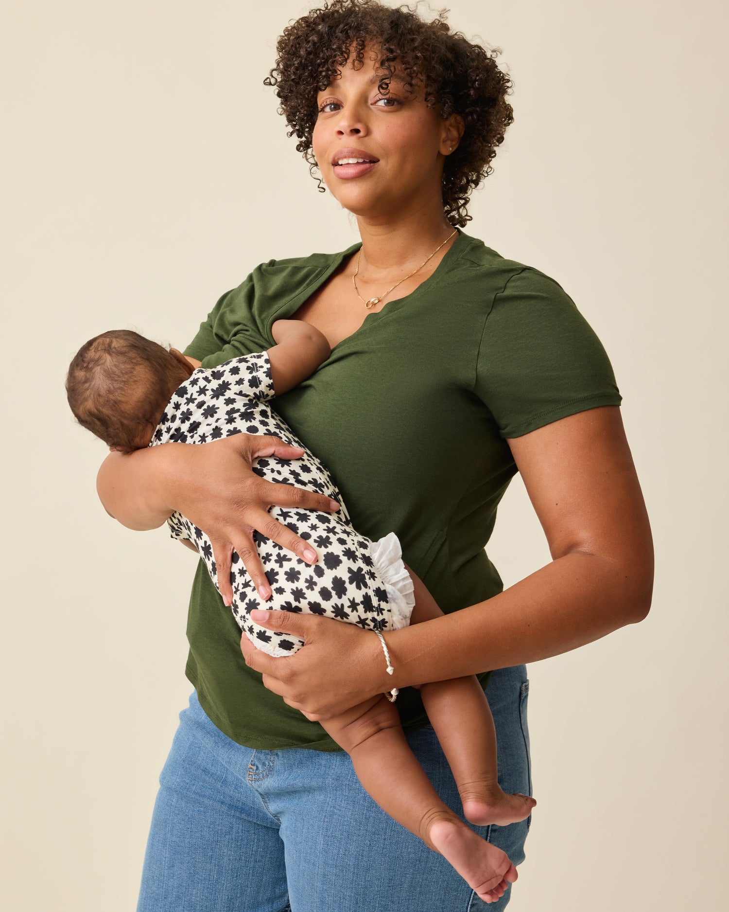 Model is breastfeeding using the front panel lift-up on the Modal V-neck Nursing T-shirt in Deep Olive on a beige background. Baby is wearing Baby Onesie in Cream Chamomiles.