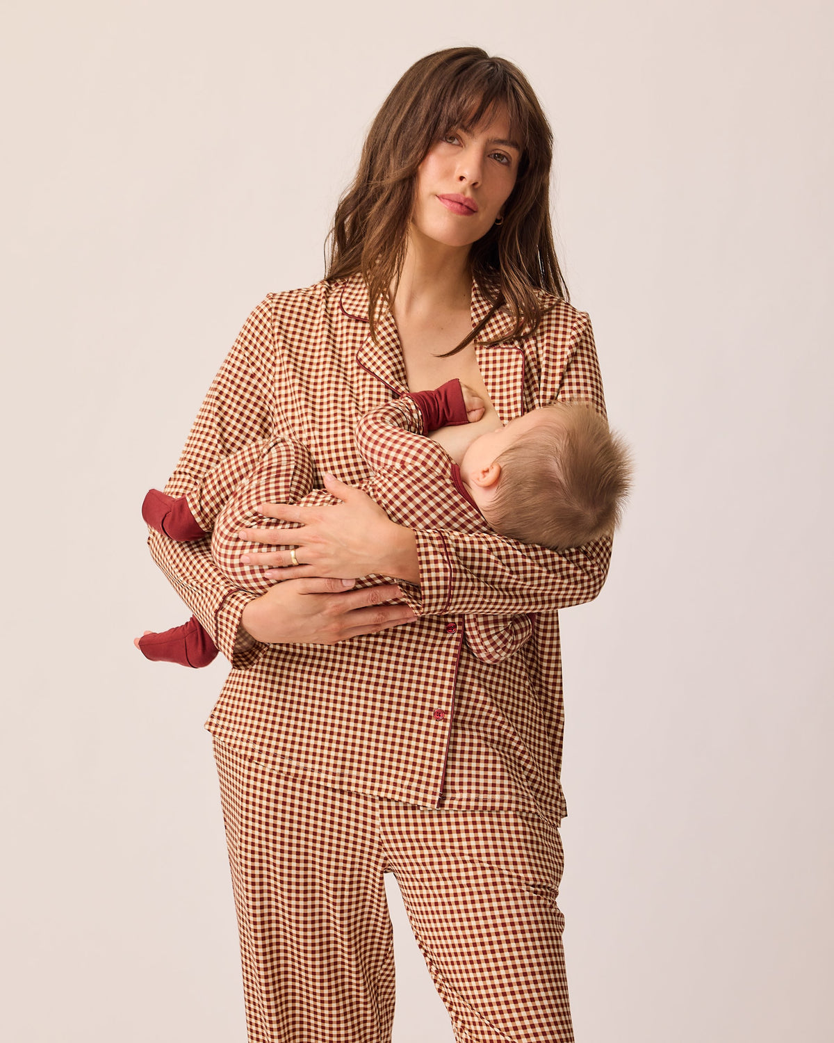 Model is holding baby while breastfeeding and wearing Clea Bamboo Long Sleeve Pajama Set in Auburn Gingham, on a beige background.