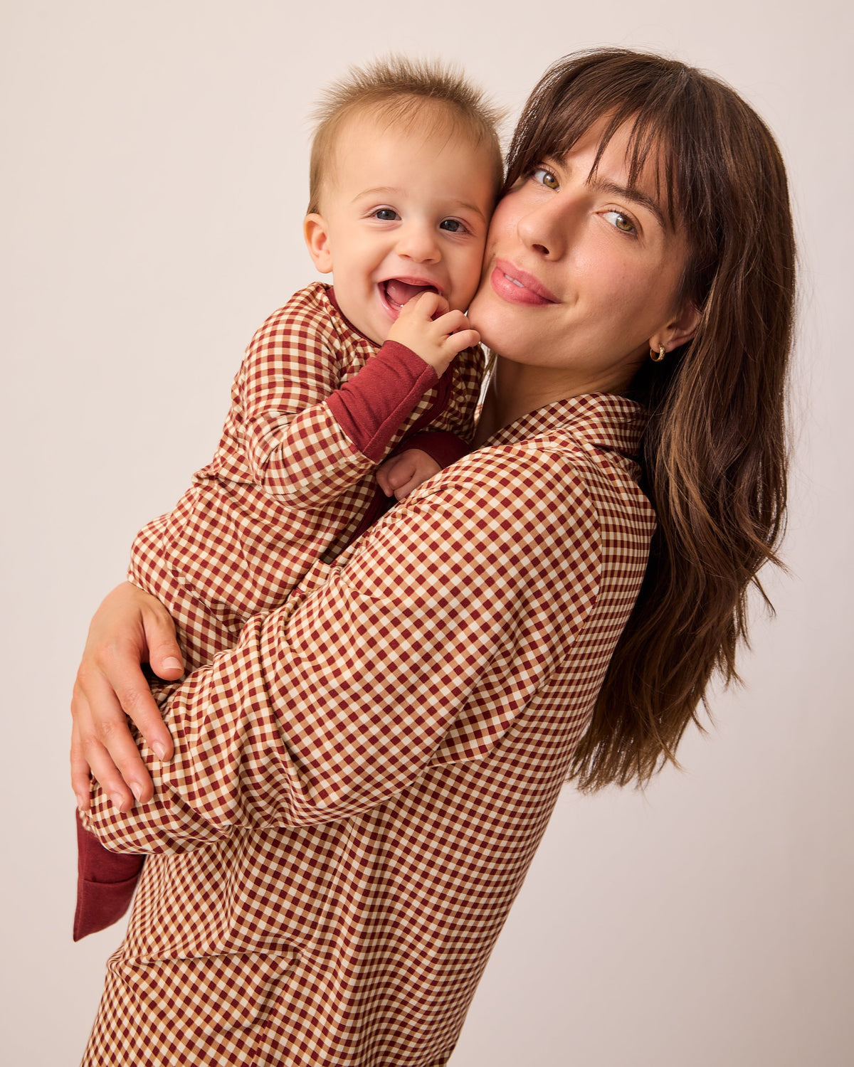 Model is holding baby in matching long sleeve onesie and wearing Clea Bamboo Long Sleeve Pajama Set in Auburn Gingham, on a beige background.