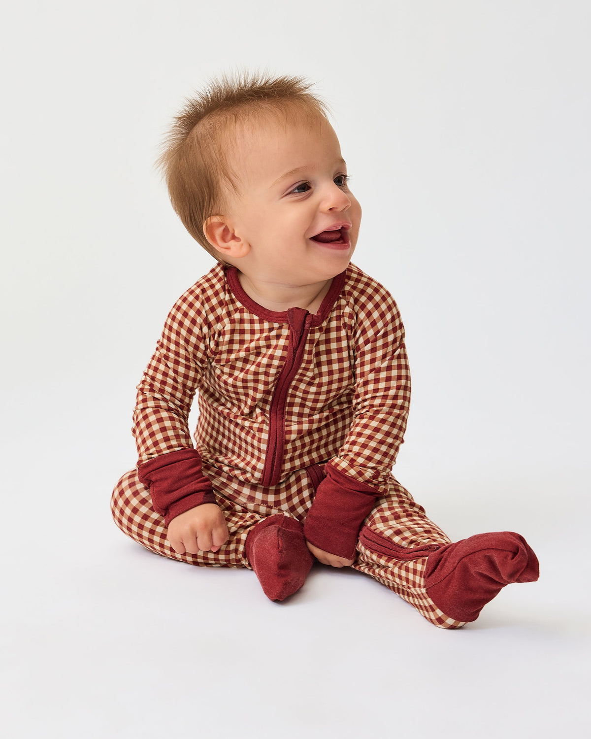 Model is sitting and wearing Bamboo Baby Pajama in Auburn Gingham on an off white background. @model_info:Shiloh is wearing 6-12 months.
