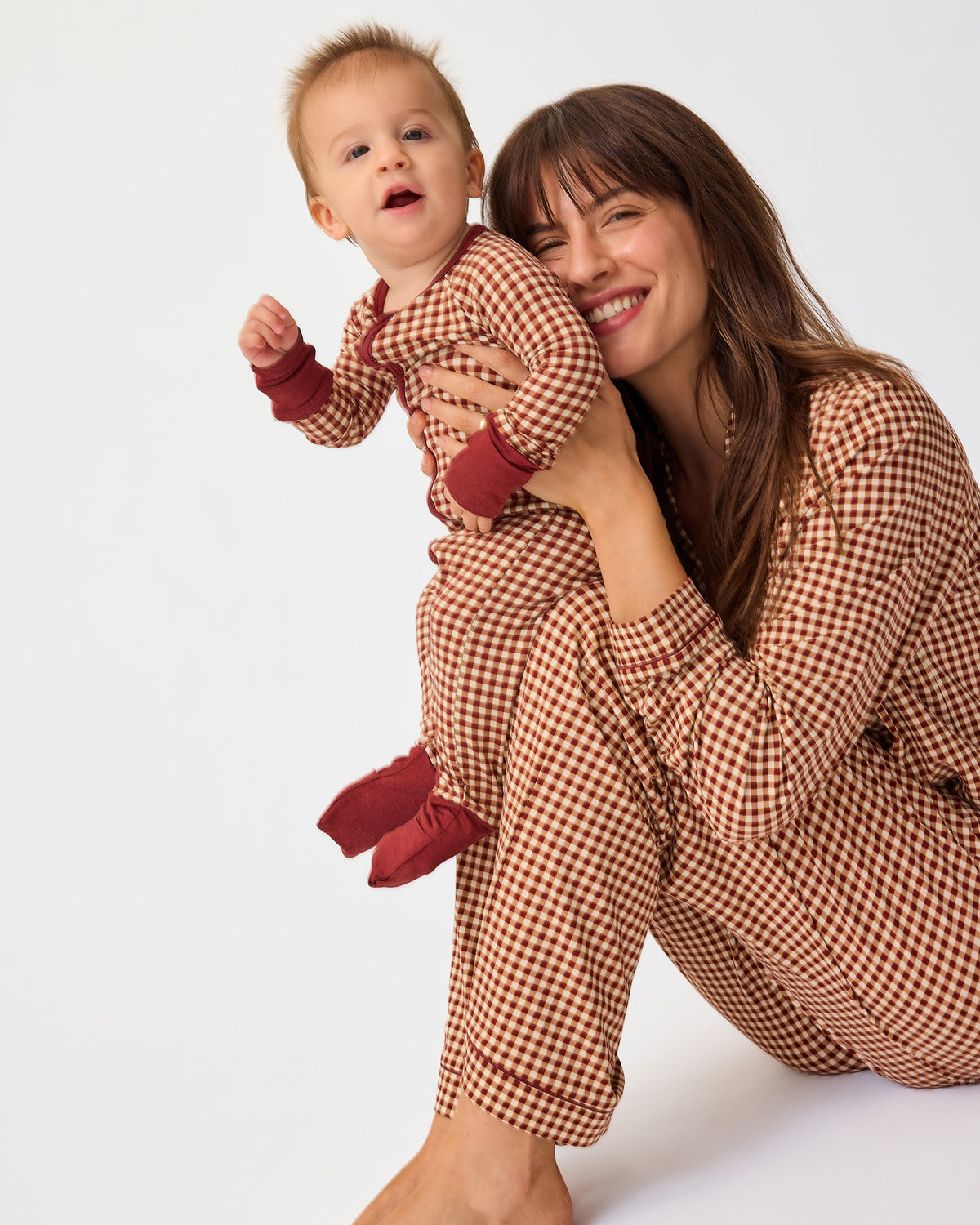 Mother is sitting and holding baby, both are wearing matching pajamas. Mother is wearing Clea Bamboo Long Sleeve Pajama Set in Auburn Gingham and Baby is wearing Bamboo Baby Pajama in Auburn Gingham on an off white background.