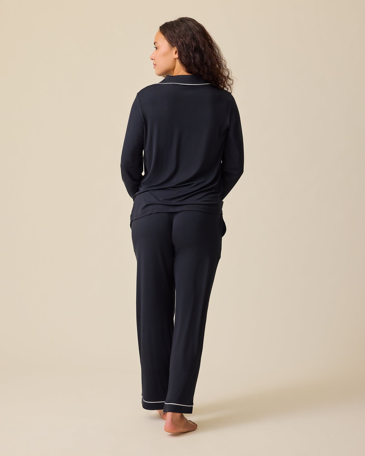 Back view of model wearing Clea Bamboo Long Sleeve Pajama Set in Black on a beige background. 