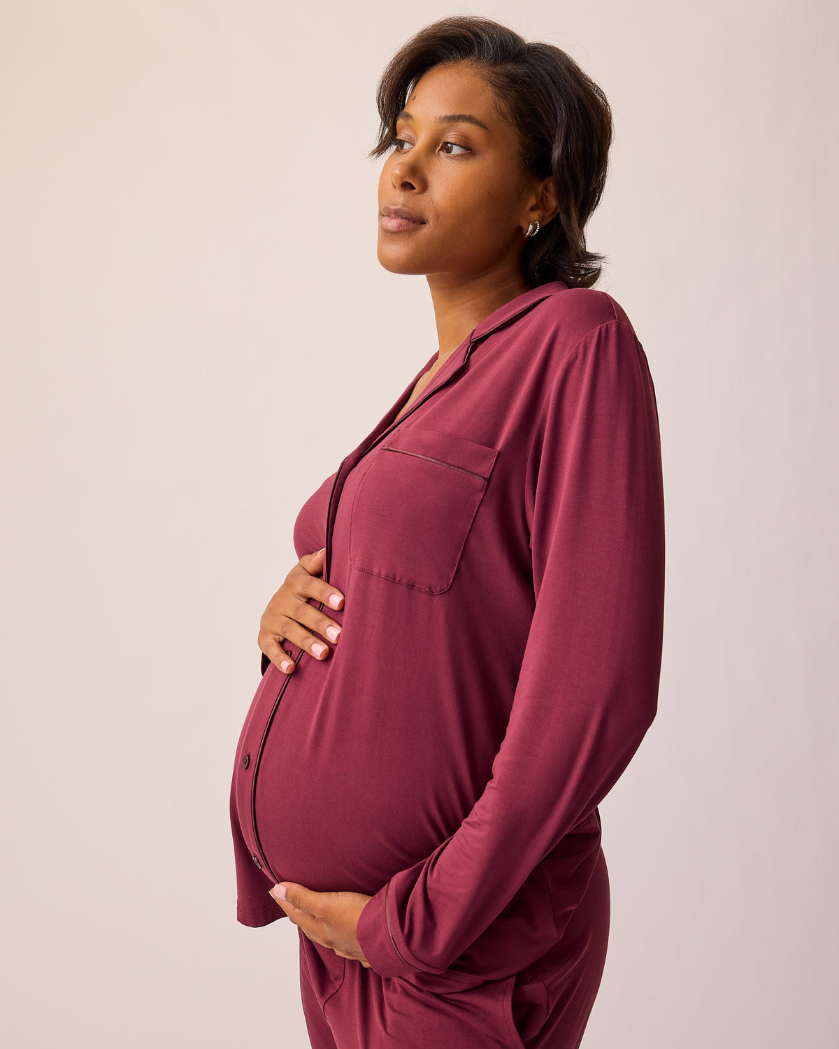 Side view of model holding baby bump and wearing Clea Bamboo Long Sleeve Pajama Set in Deep Burgundy on a beige background.
