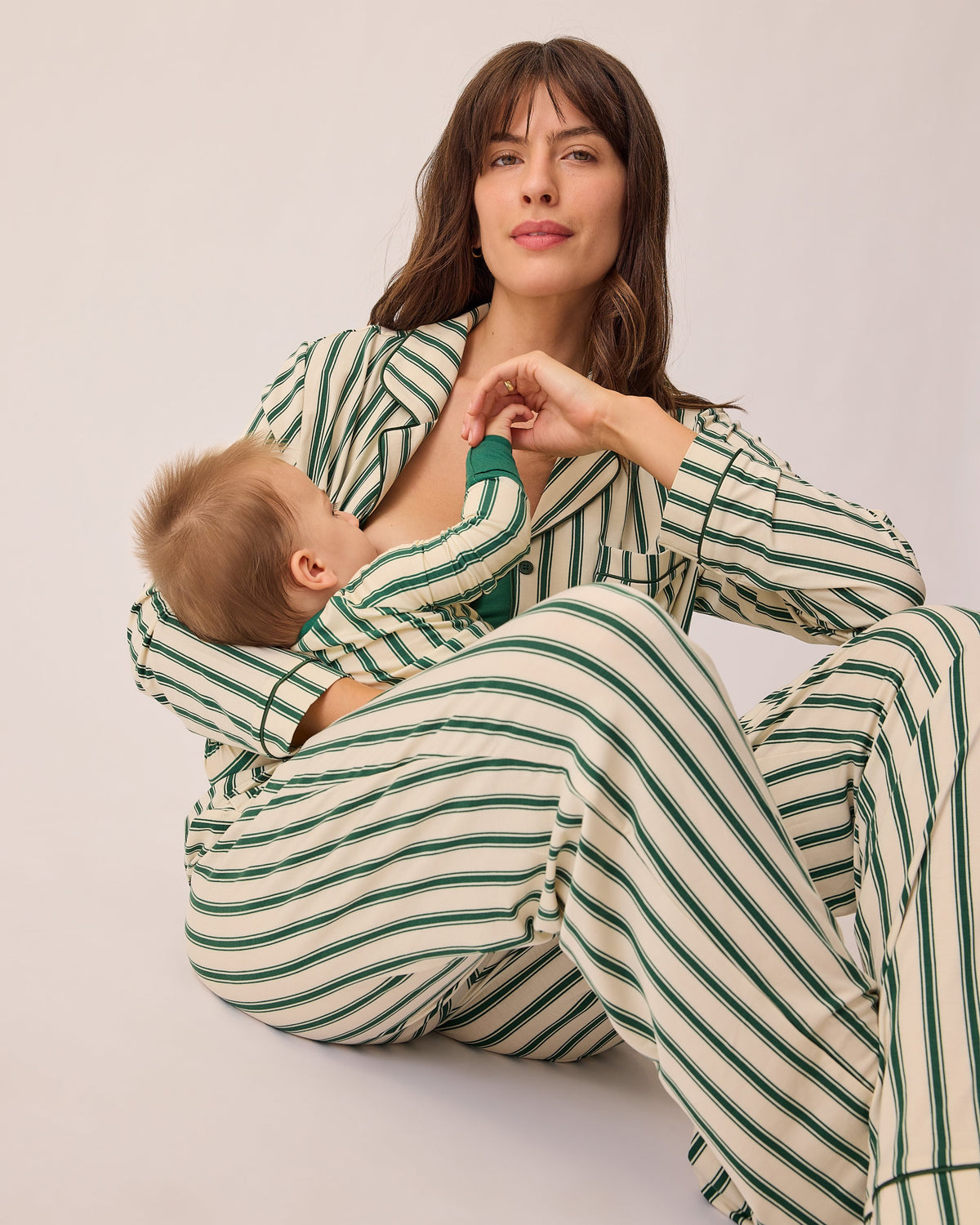Model is holding baby in matching long sleeve onesie and breastfeeding while sitting on floor and wearing Clea Bamboo Long Sleeve Pajama Set in Green Candy Stripe, on a beige background.