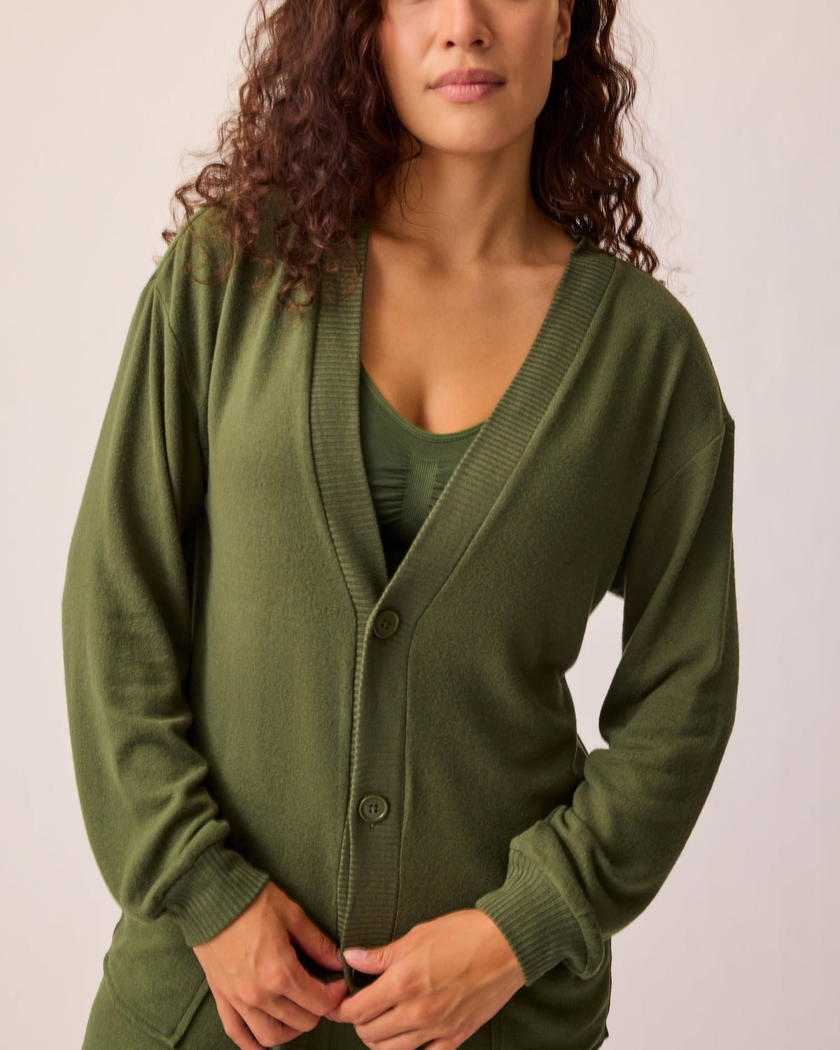 Model is buttoning CozyKnit Long Cardigan in Deep Olive, with Simply Sublime® Nursing Bra in Deep Olive, and CozyKnit Wide Leg Lounge Pant in Deep Olive on a beige background. 