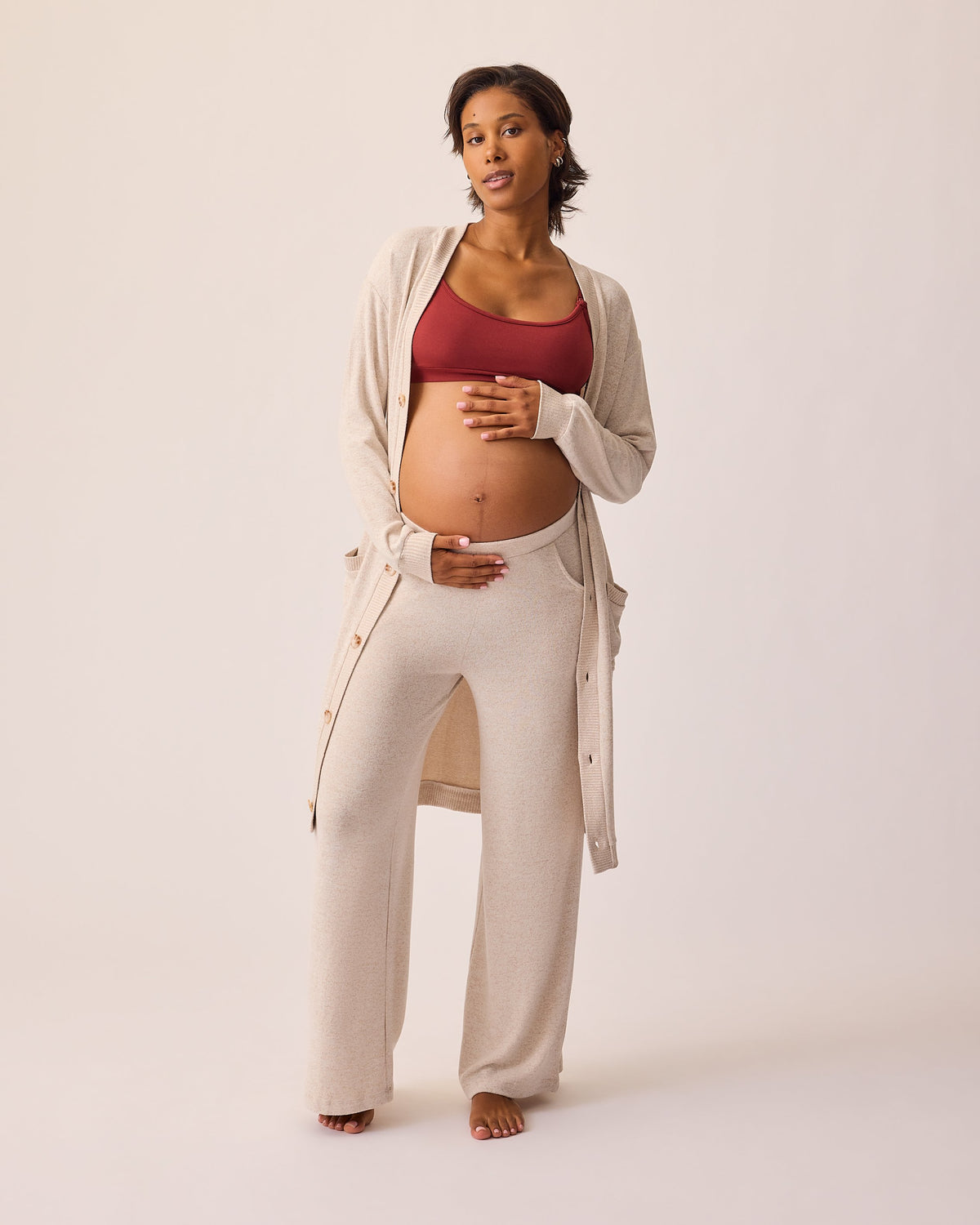 Model is wearing CozyKnit Wide Leg Lounge Pant in Oat, and CozyKnit Long Cardigan in Oat, and French Terry Maternity & Nursing Bralette in Deep Burgundy on a beige background.