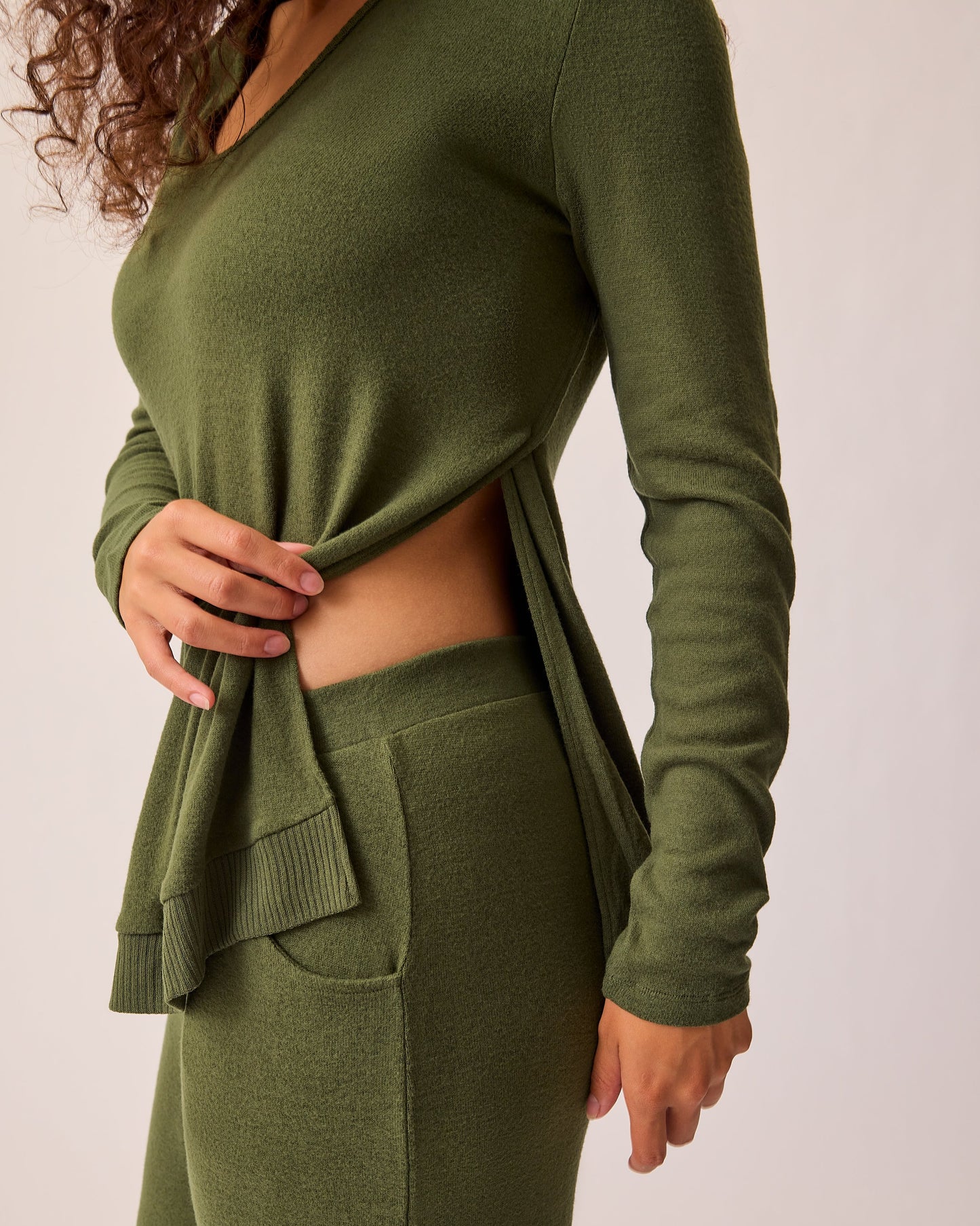 Model is lifting front from side to show ease of nursing on the CozyKnit Maternity & Nursing V-neck Top in Deep Olive on a beige background. 