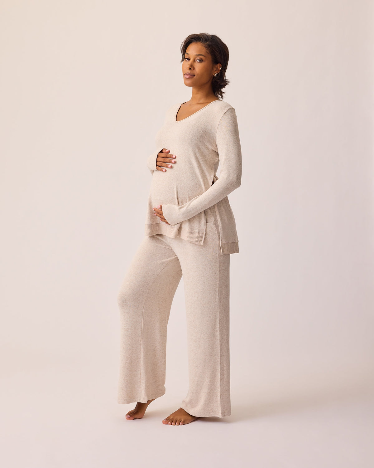 Angled view of model wearing CozyKnit Maternity & Nursing V-neck Top in Oat and CozyKnit Wide Leg Lounge Pant in Oat on a beige background.