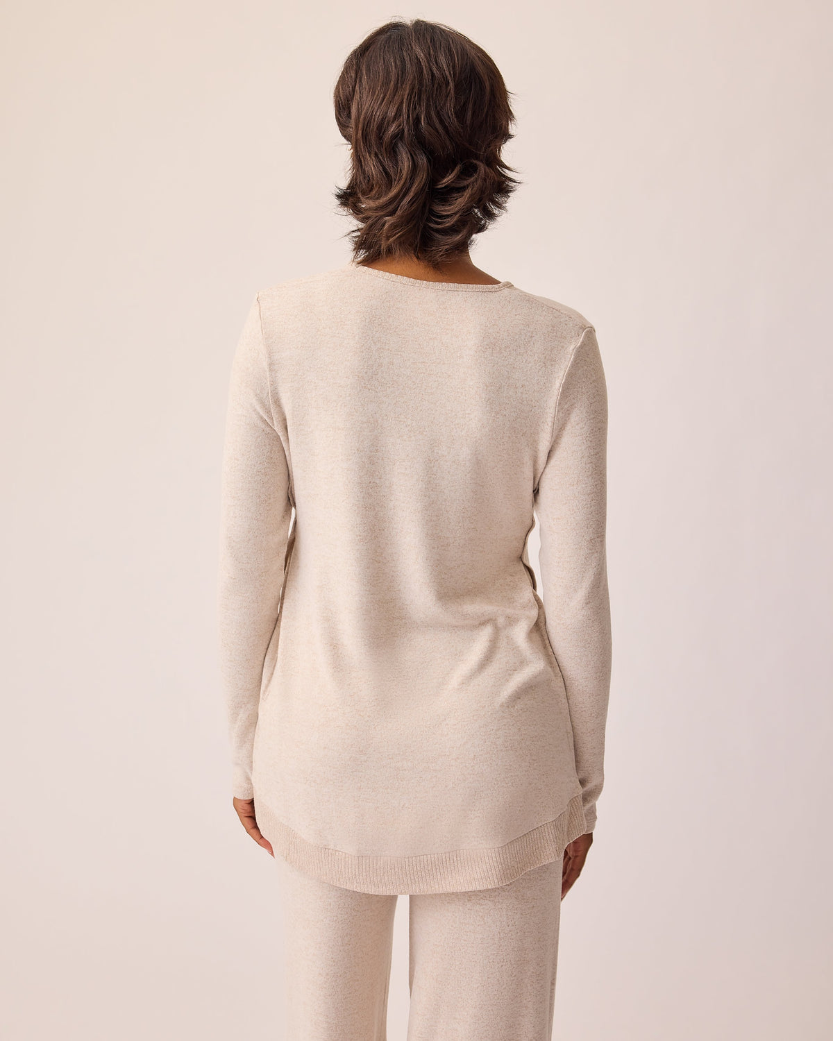 Back view of the model wearing CozyKnit Maternity & Nursing V-neck Top in Oat and CozyKnit Wide Leg Lounge Pant in Oat on a beige background.