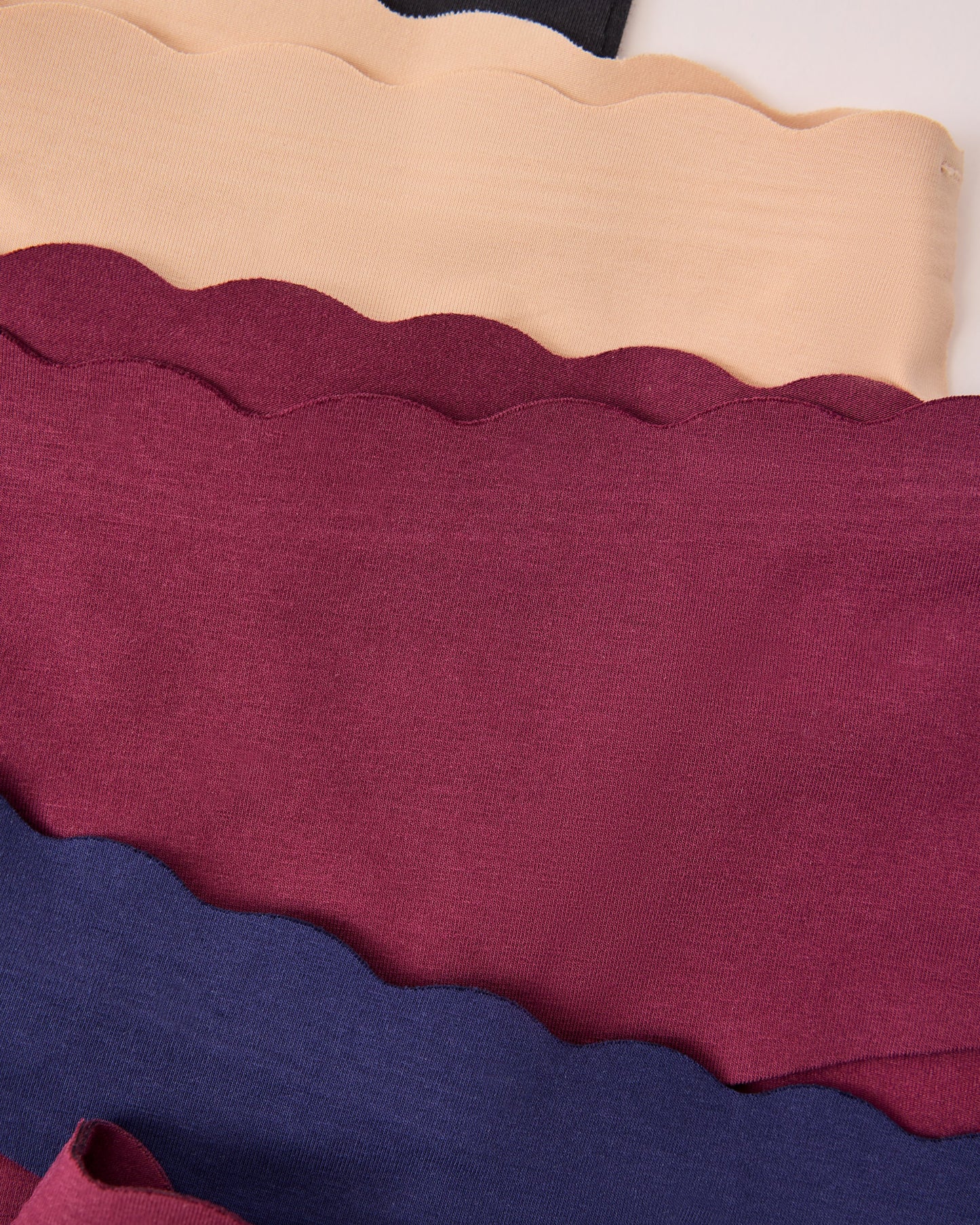 Four scalloped-edge fabric bands of the BareBond™ Cotton Maternity & Postpartum Underwear in Black, Sand, Garnet and Coastal Navy on a beige background.