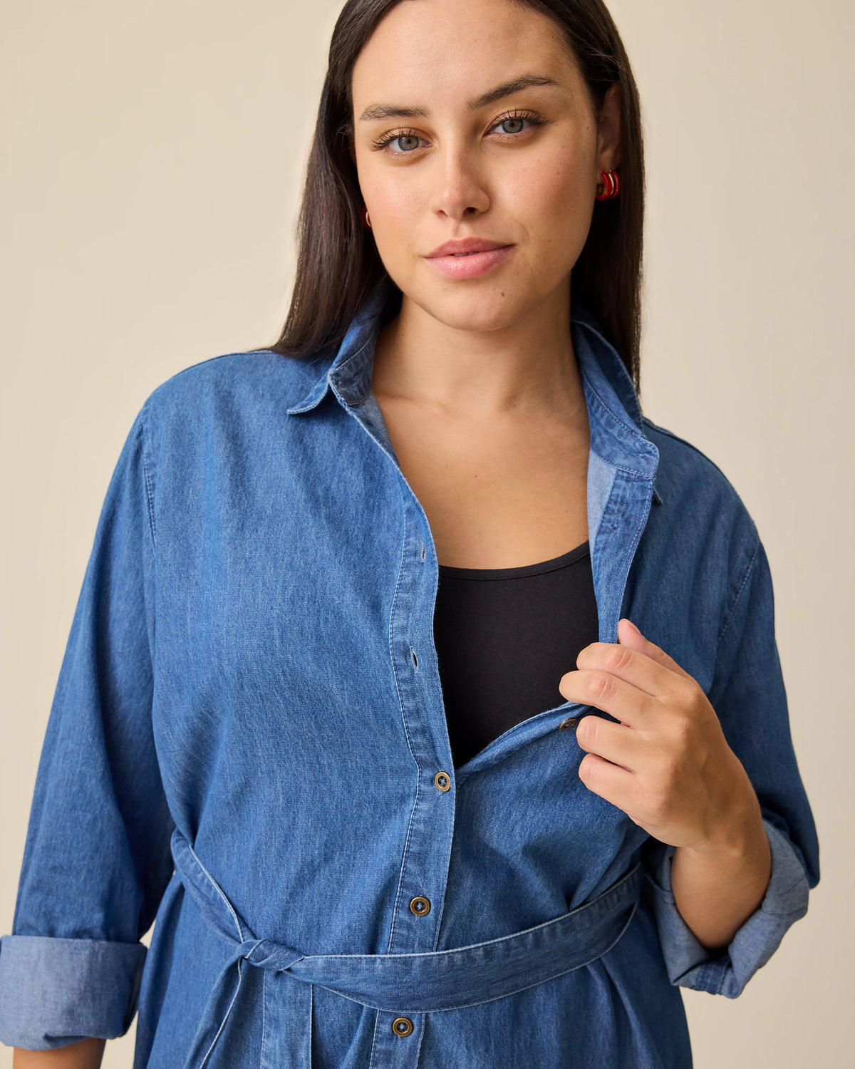 Model pulls unbuttoned front open to demonstrate nursing access on the Denim Maternity & Nursing Shirt Dress in Dark Wash on a beige background.