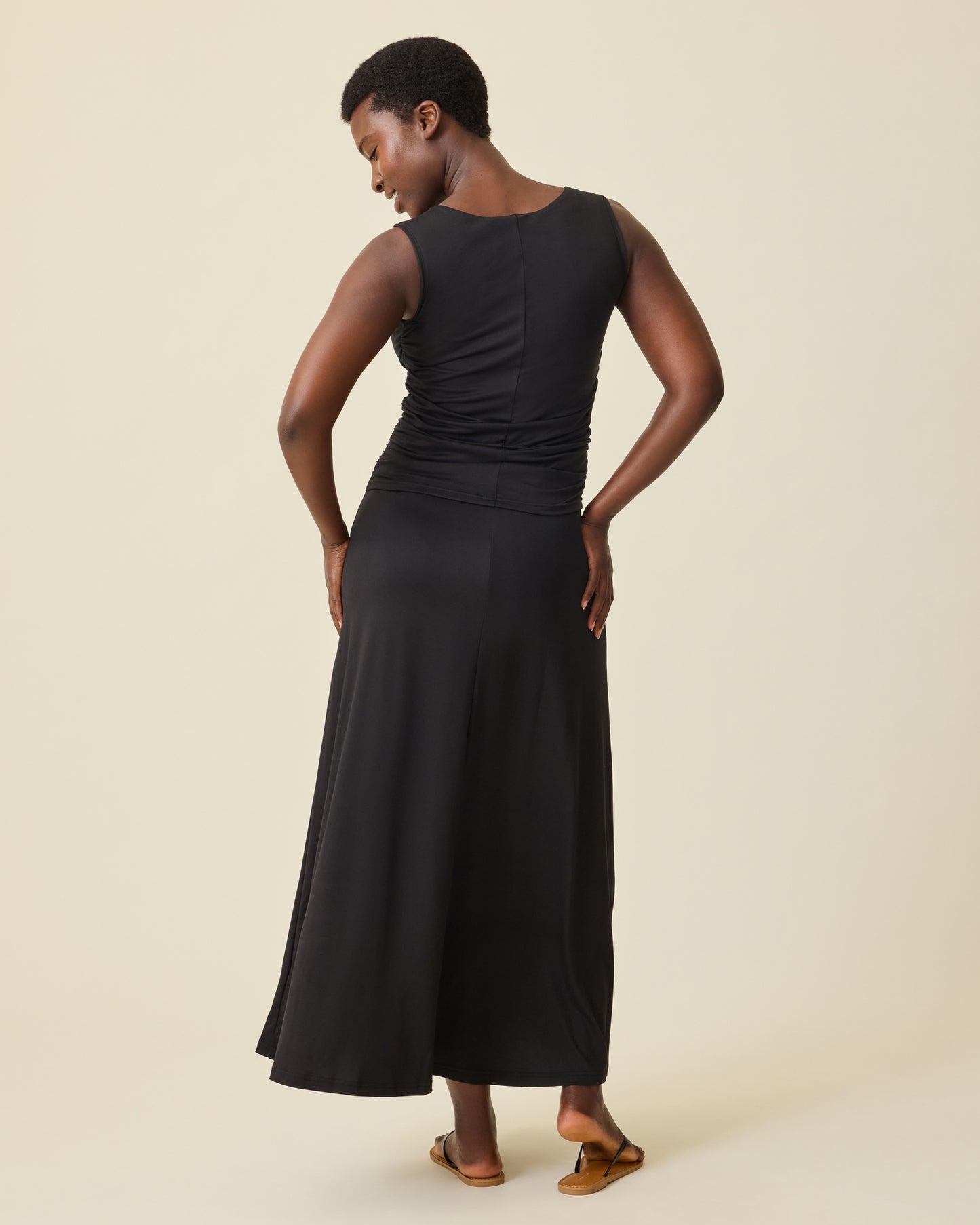 Back view of pregnant model wearing Drop-Waist Maternity & Nursing Maxi Dress in Black on a beige background