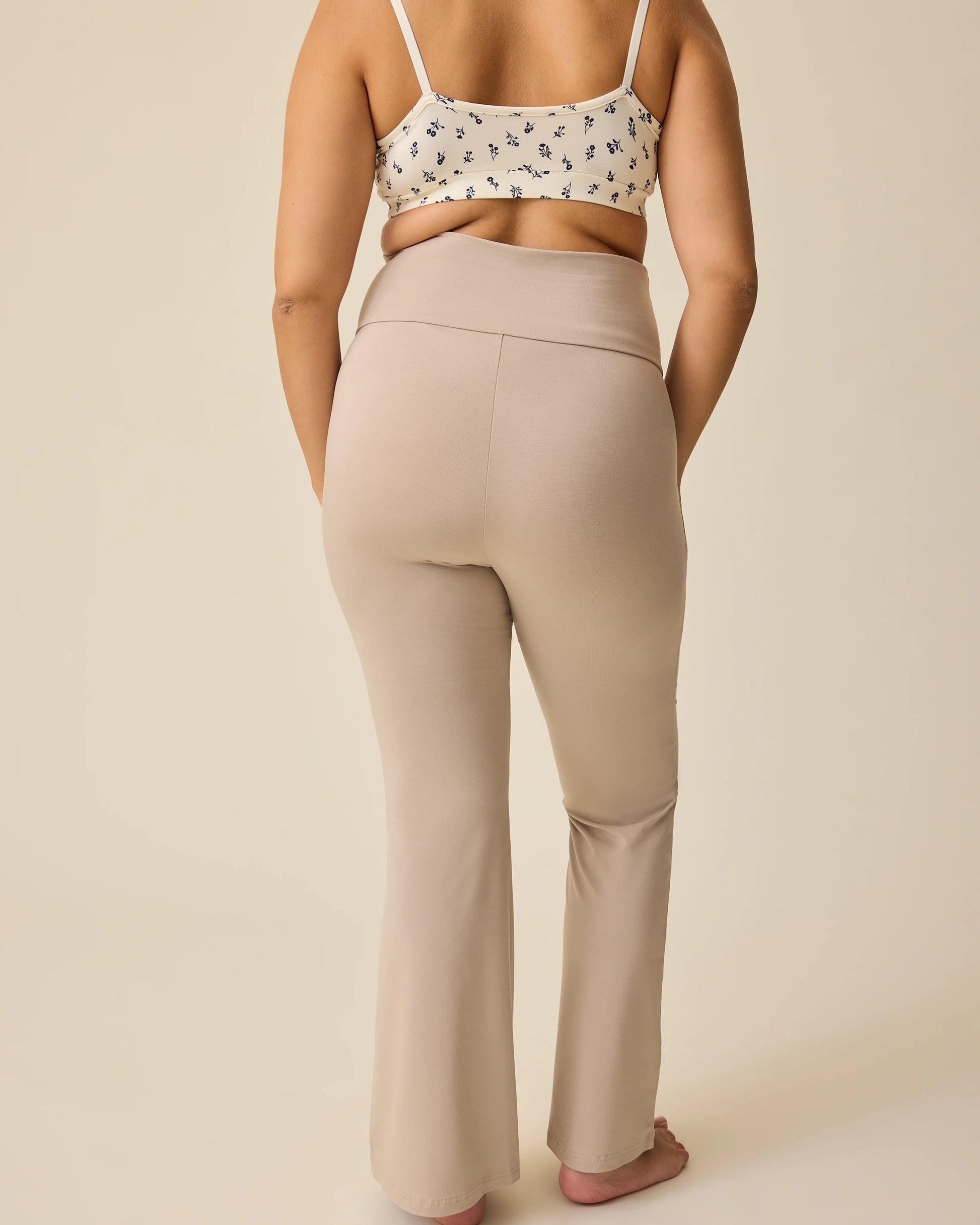 Back view of model wearing French Terry Racerback Nursing & Sleep Bra in Ivory Wildflower and Bamboo Flex Maternity & Postpartum Flare Pant in Sandstone on a beige background.