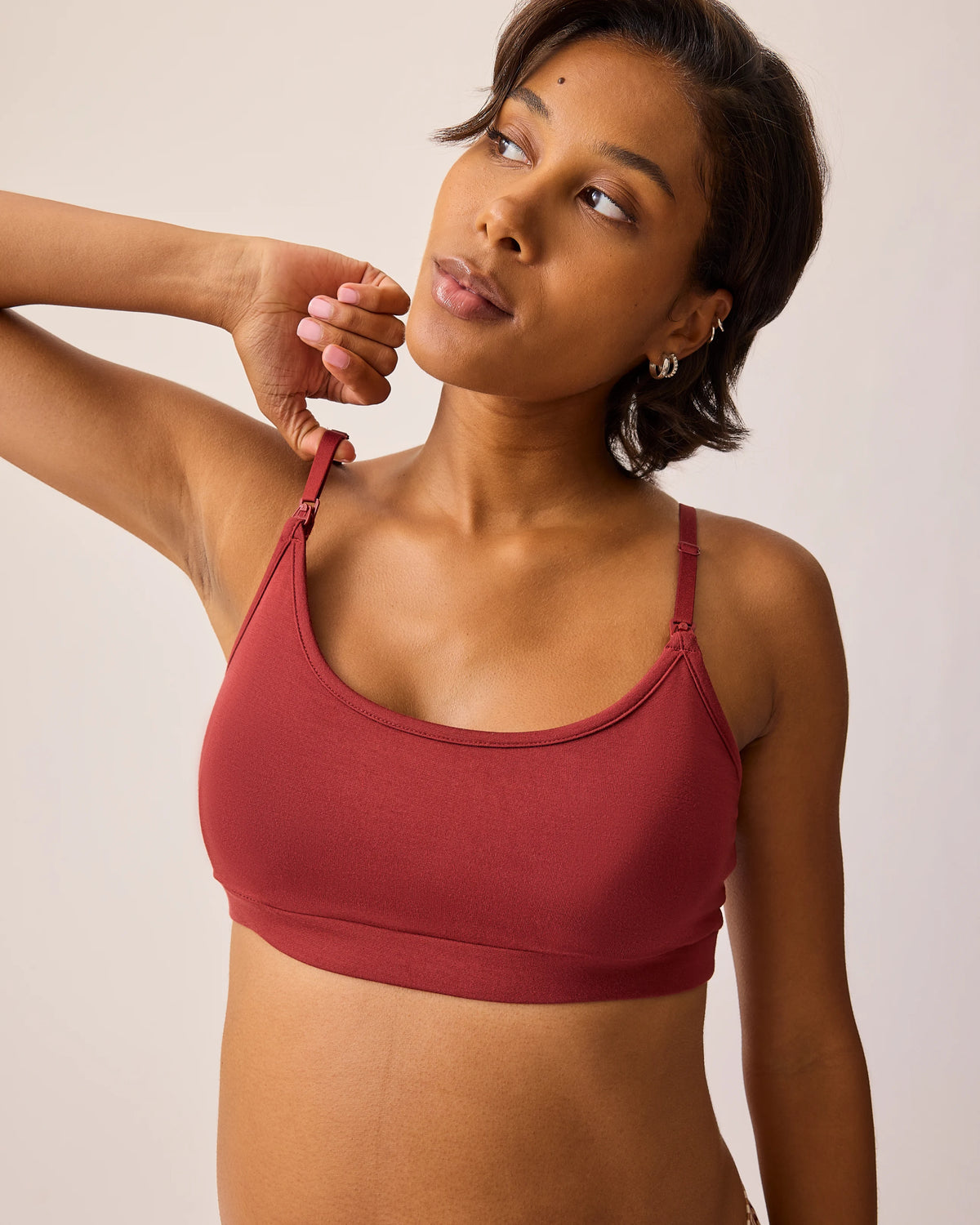Model is wearing the French Terry Maternity & Nursing Bralette in Auburn while stretching the strap on one side, on a beige background. @model_info:Ale is wearing a Medium.