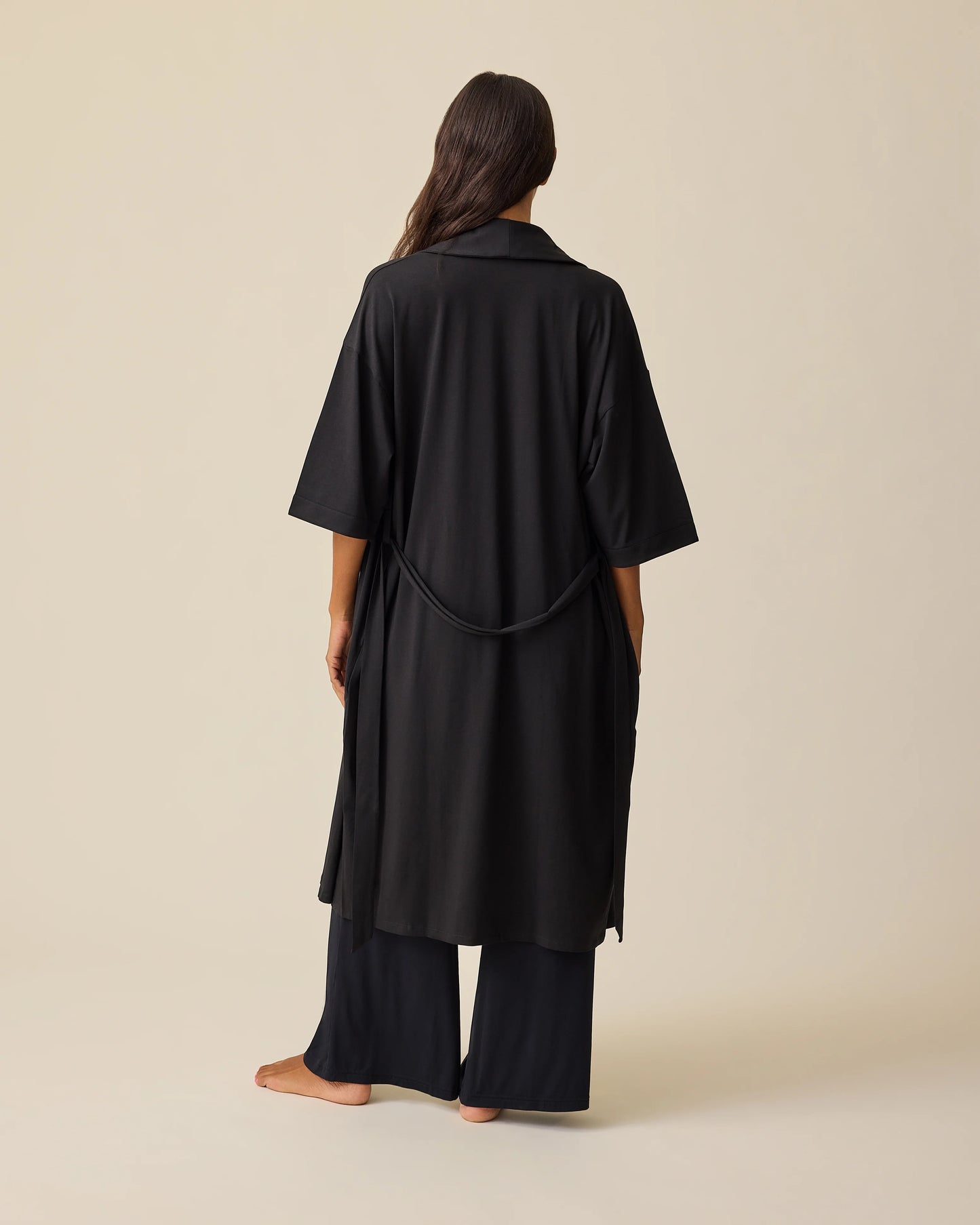 Back view of model wearing The Restore Cotton Blend Robe in Black on a beige background.