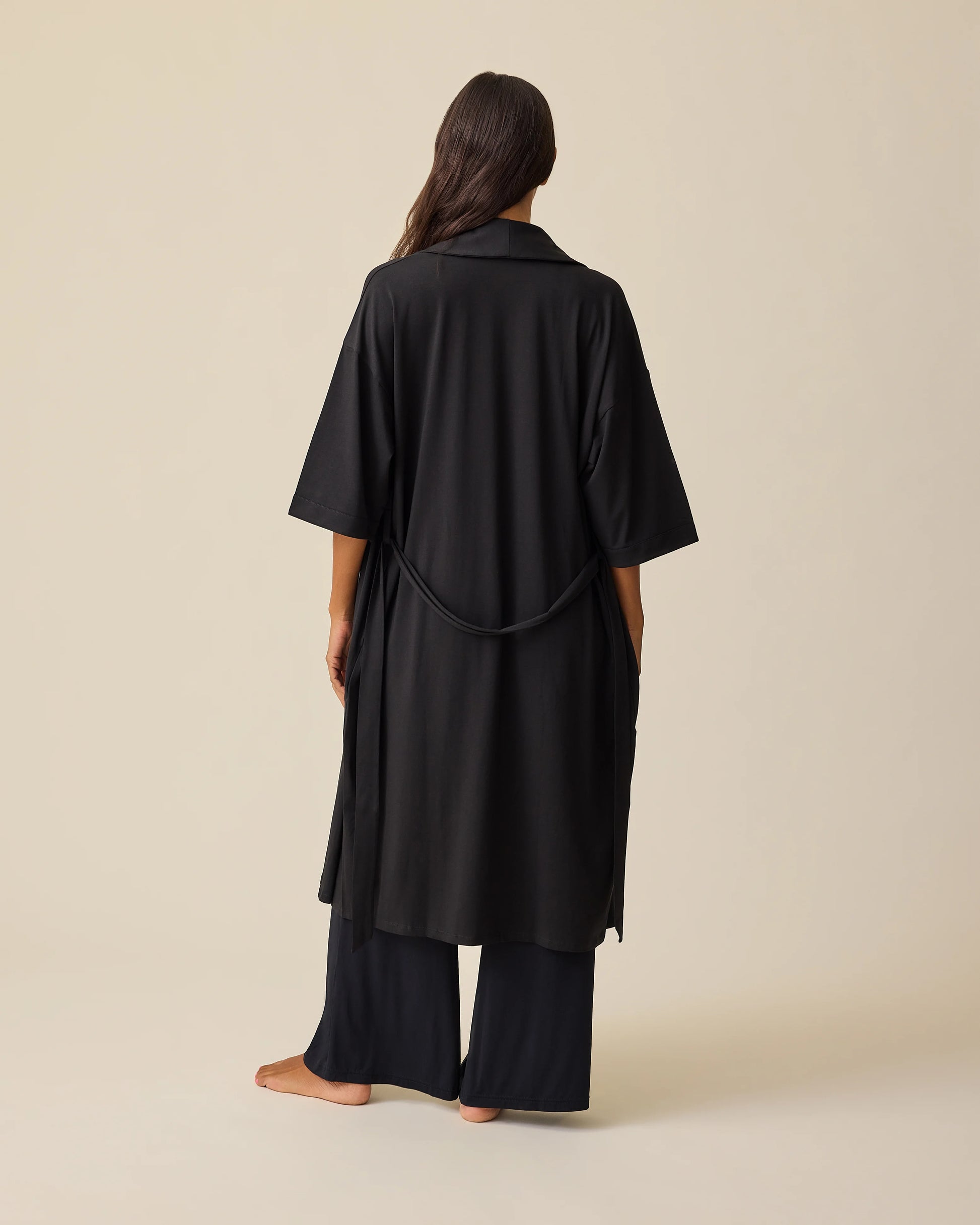 Back view of model wearing The Restore Cotton Blend Robe in Black on a beige background.