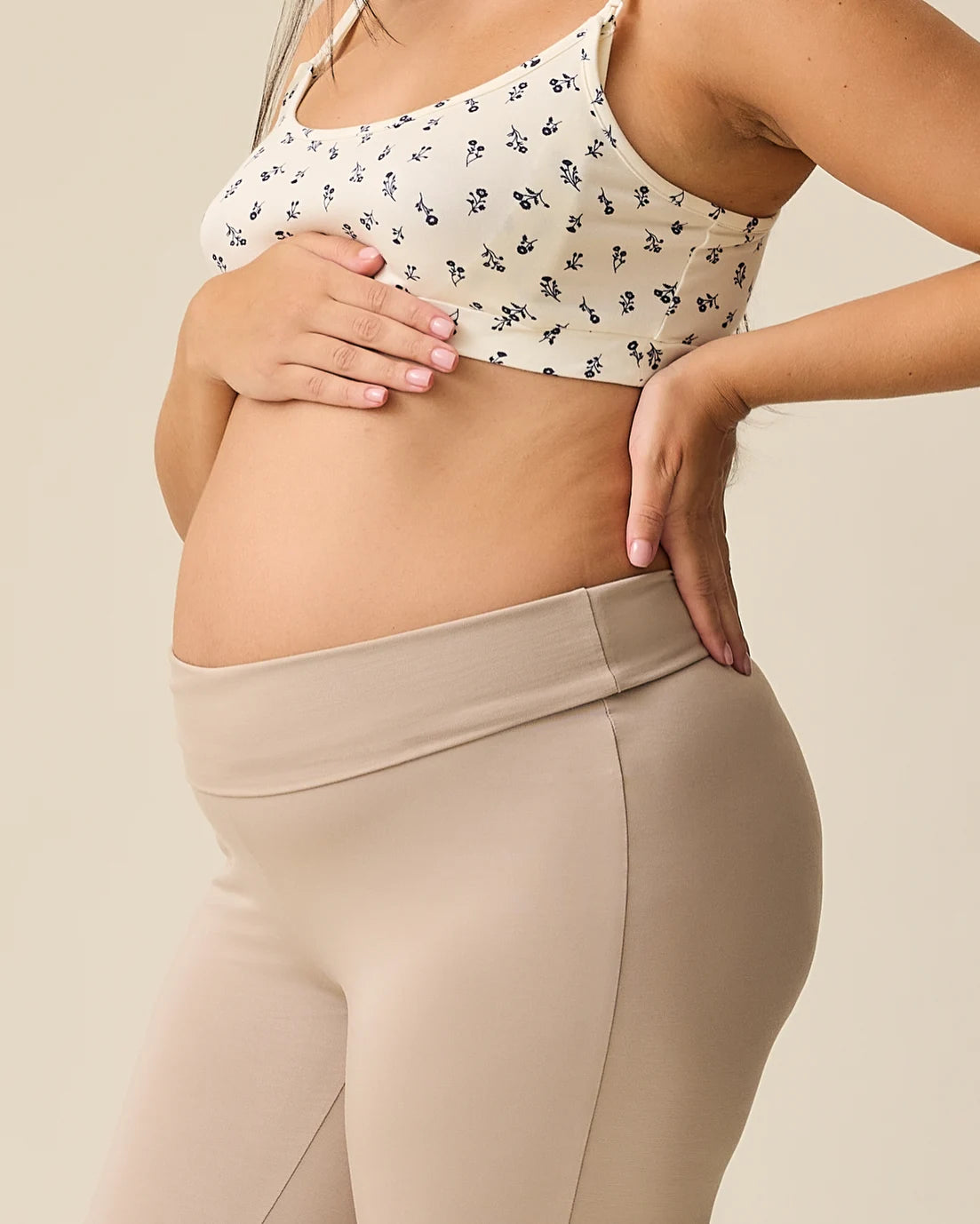 Side view of model wearing French Terry Racerback Nursing & Sleep Bra in Ivory Wildflower and Bamboo Flex Maternity & Postpartum Flare Pant in Sandstone with waist folded on a beige background.