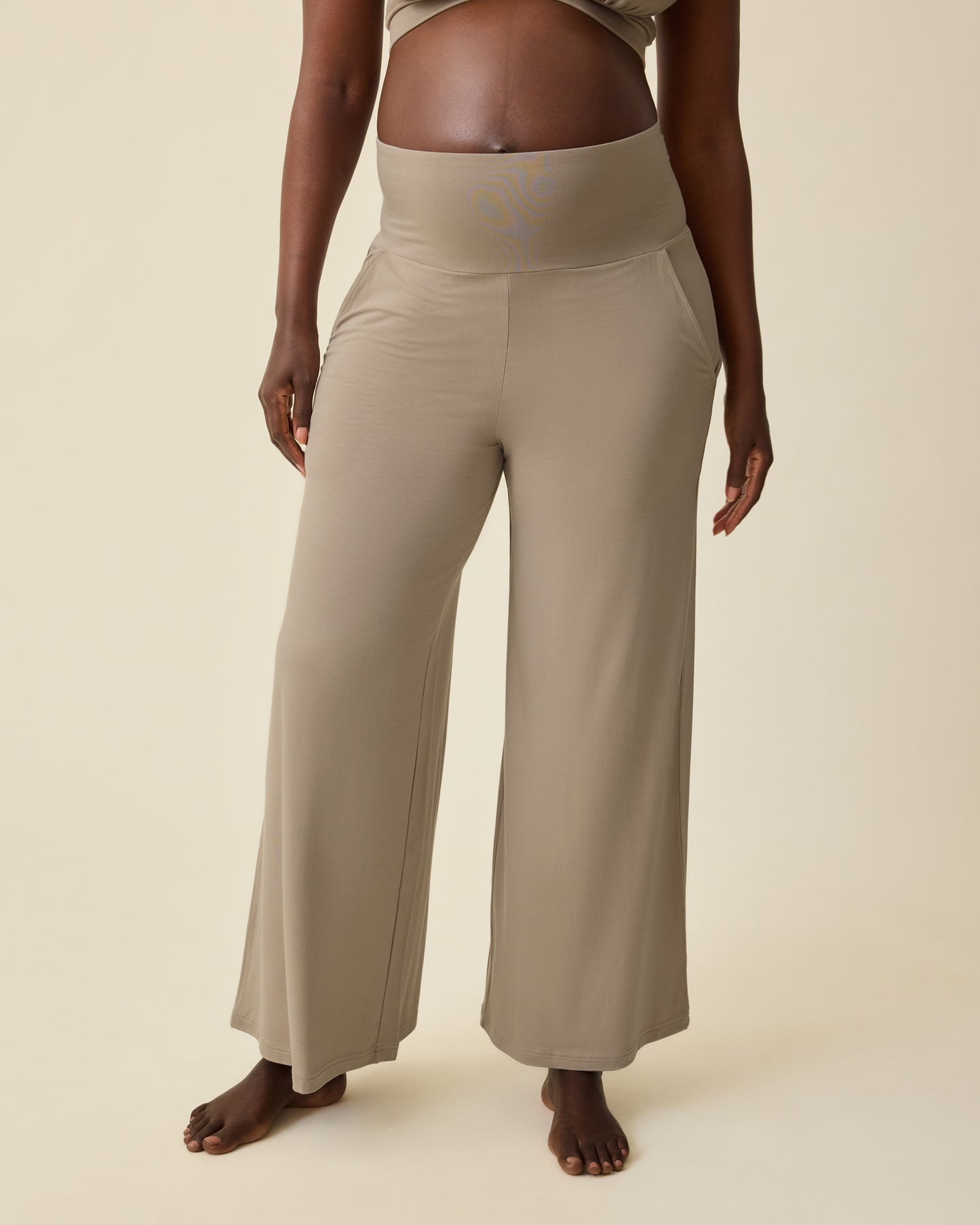 Pregnant model is wearing Bamboo Wide Leg Maternity & Postpartum Lounge Pant in Sandstone on a beige background.