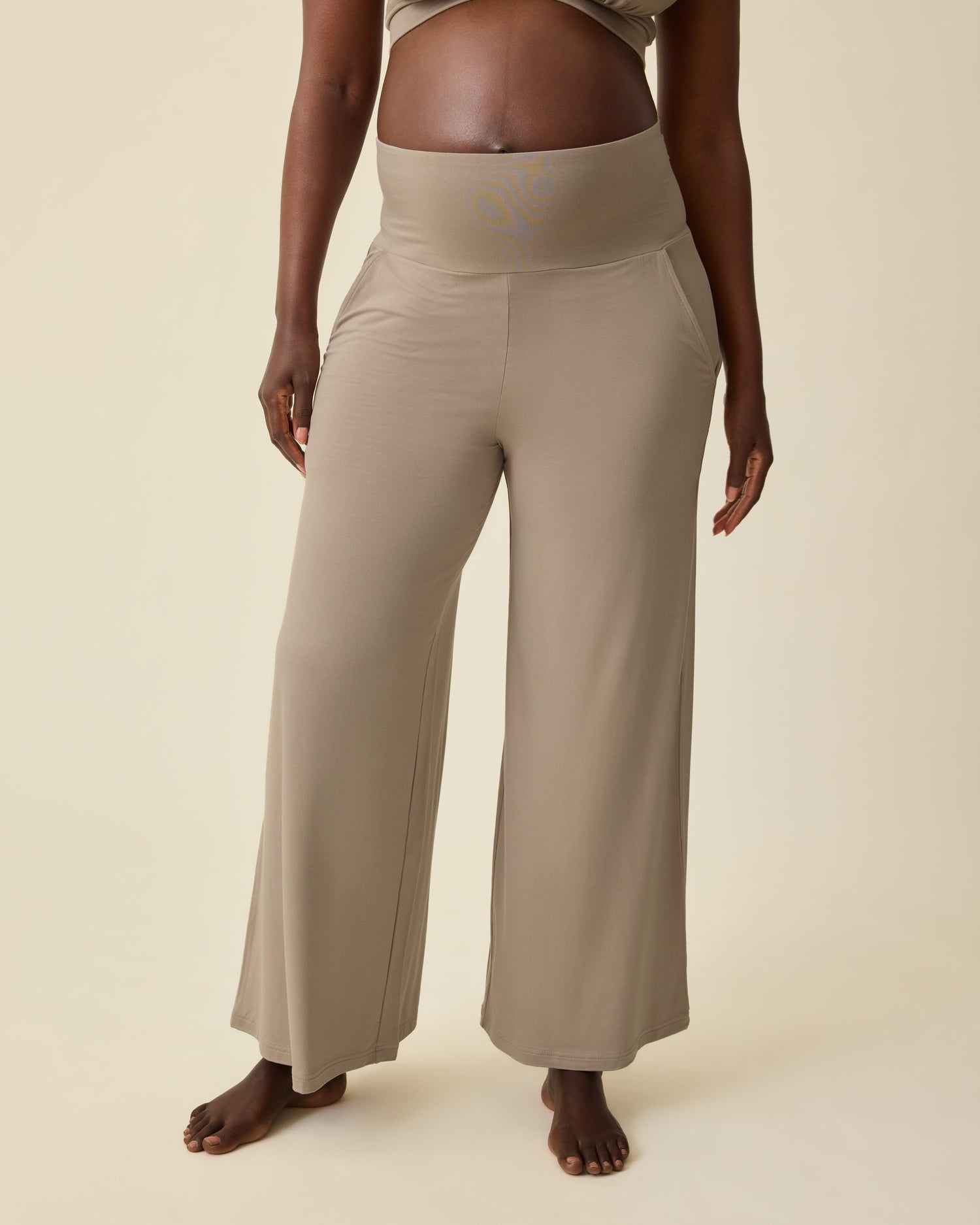 Pregnant model is wearing Bamboo Wide Leg Maternity & Postpartum Lounge Pant in Sandstone on a beige background.