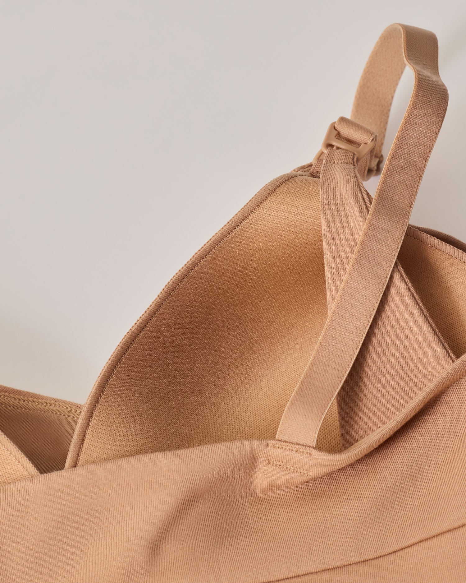 The back view of SoftStretch Cotton Nursing Bra in Buff, laying on surface. Soft material and color, support straps and nursing clasps. On a beige background.